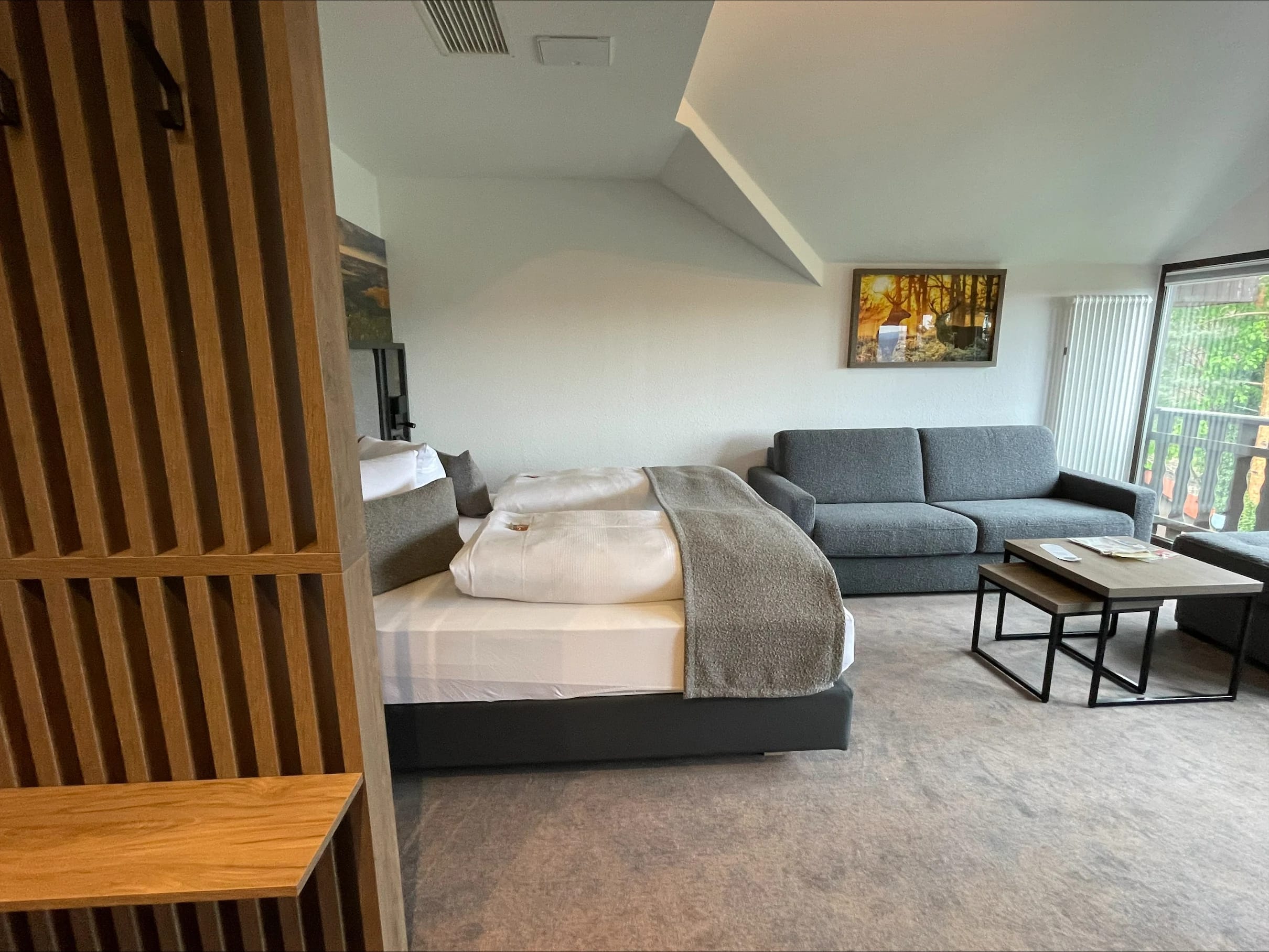 Zimmer mit Doppelbett, grauem Sofa und Dachschräge, daneben ein Raumteiler aus Holzlamellen