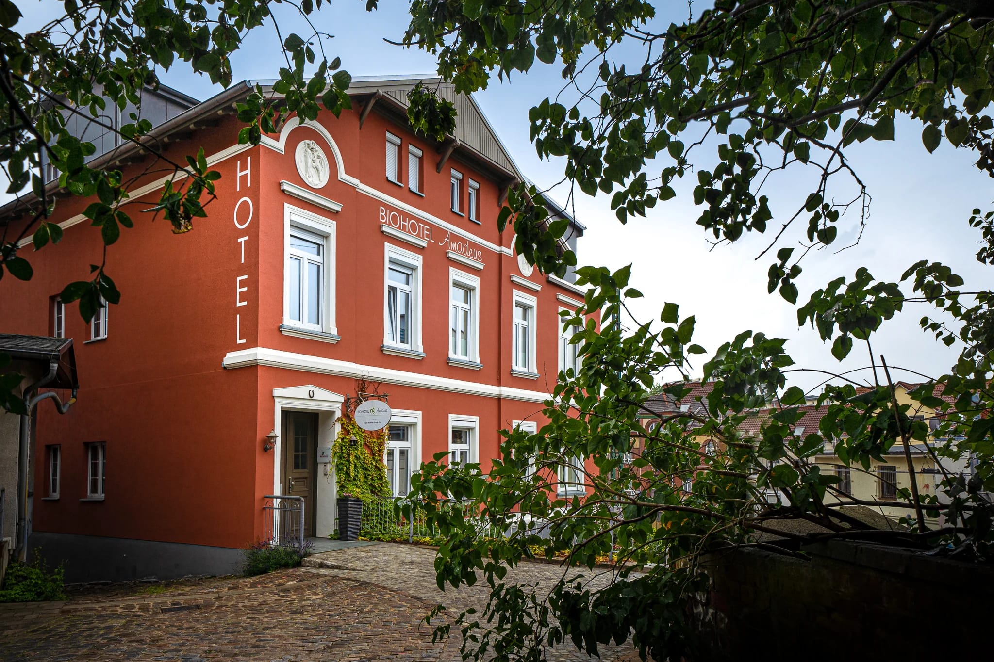 Rotes Hotelgebäude am Kopfsteinpflasterplatz, von grünen Zweigen eingerahmt