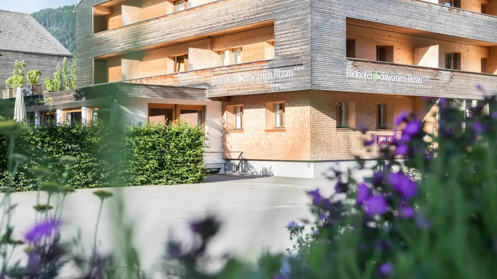 Außenansicht des Hotels im Sommer, Holzfassade mit Balkonen hinter blühenden Stauden