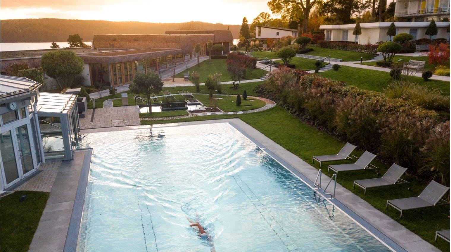 Beheiztes Freibad mit Liegestühlen im Abendlicht, dahinter die Gartenanlage und der See