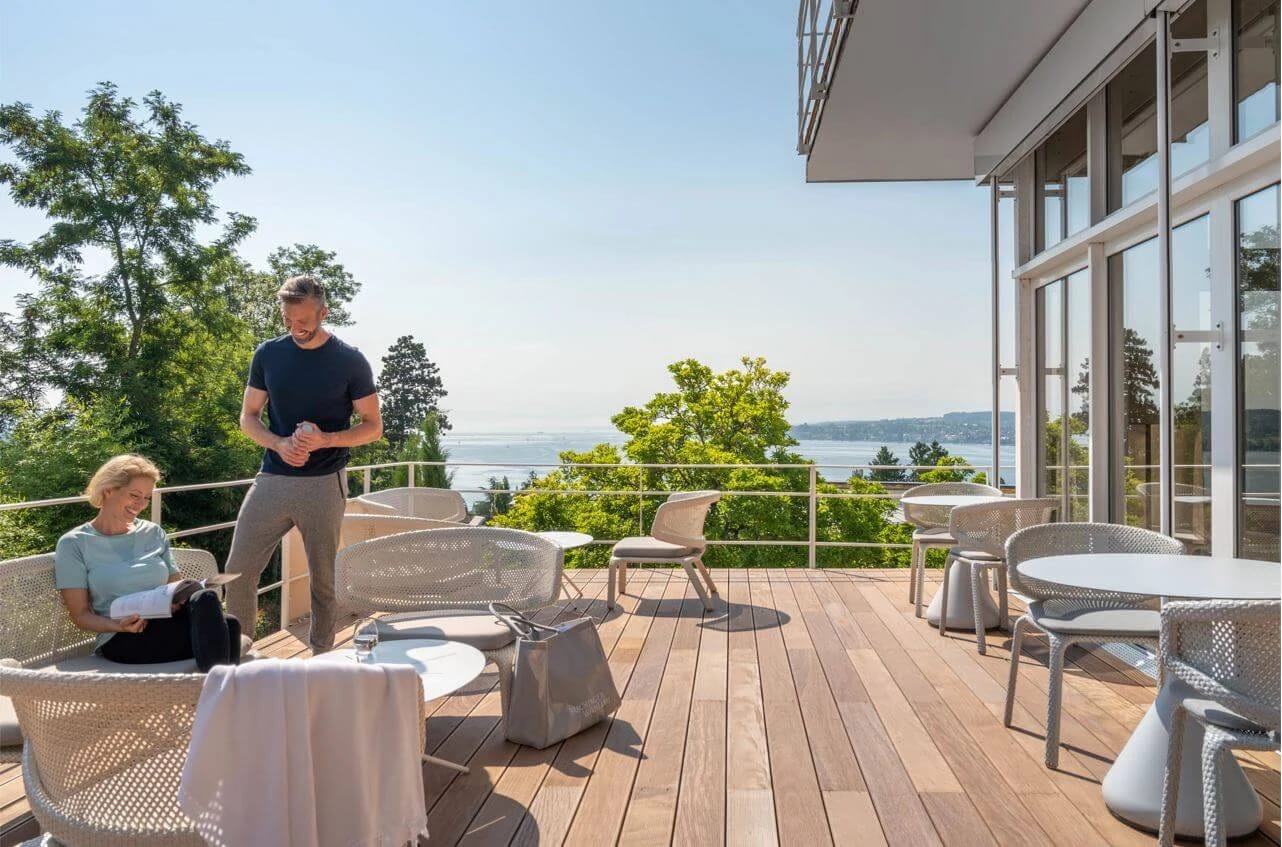 Holzterrasse mit Tischen und Korbsesseln vor der Glasfront des Haupthauses, mit Blick über Bäume auf den See