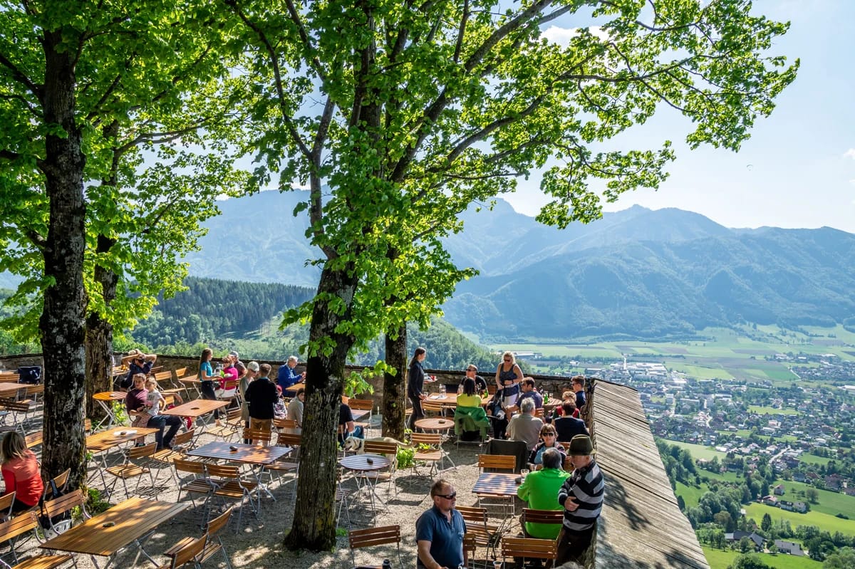 Burgterrasse unter großen Bäumen mit Sitzbänken und Blick über das Tal auf die Berge