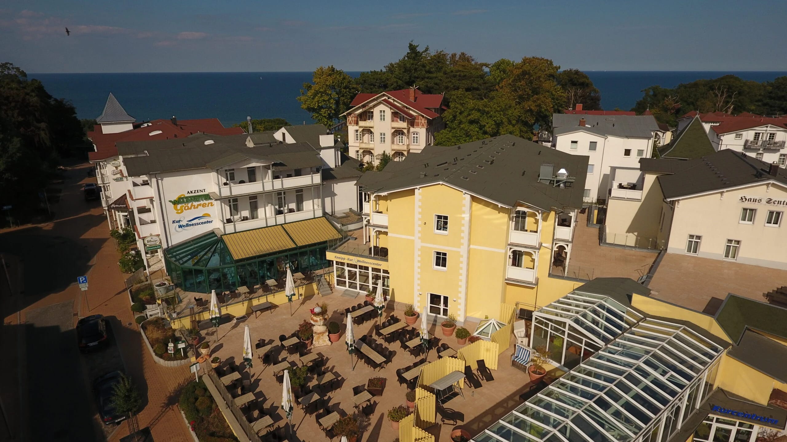 Luftbild des Hotels mit gelbem Haupthaus, Terrasse mit Sonnenschirmen und Glaspavillon, im Hintergrund die Ostsee