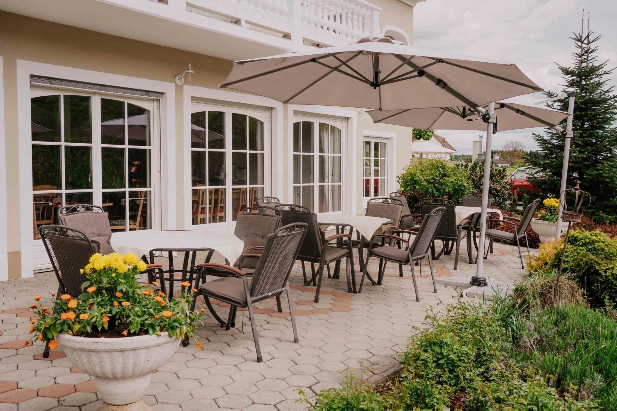 Terrasse mit Tischen, Stühlen und Sonnenschirm vor den Bogenfenstern des Hauses