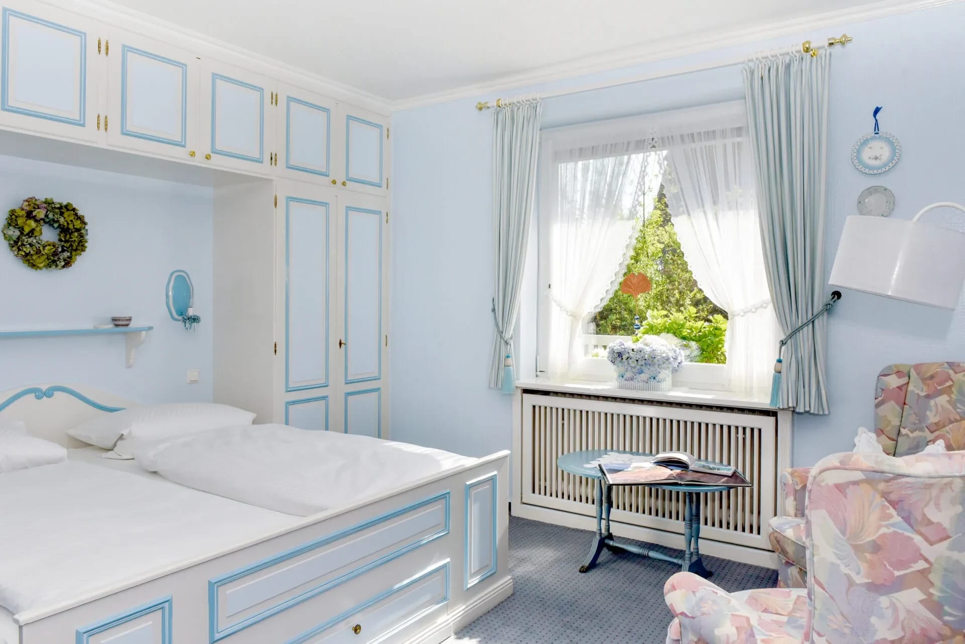 Hellblau eingerichtetes Doppelzimmer mit weißem Bett, Schrankwand und Sessel am Fenster