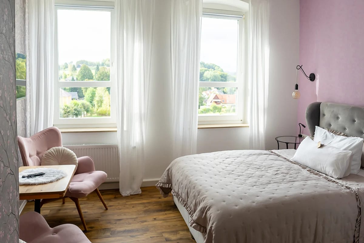 Doppelzimmer mit rosa Sesseln und Blick durch zwei Fenster auf den Ort