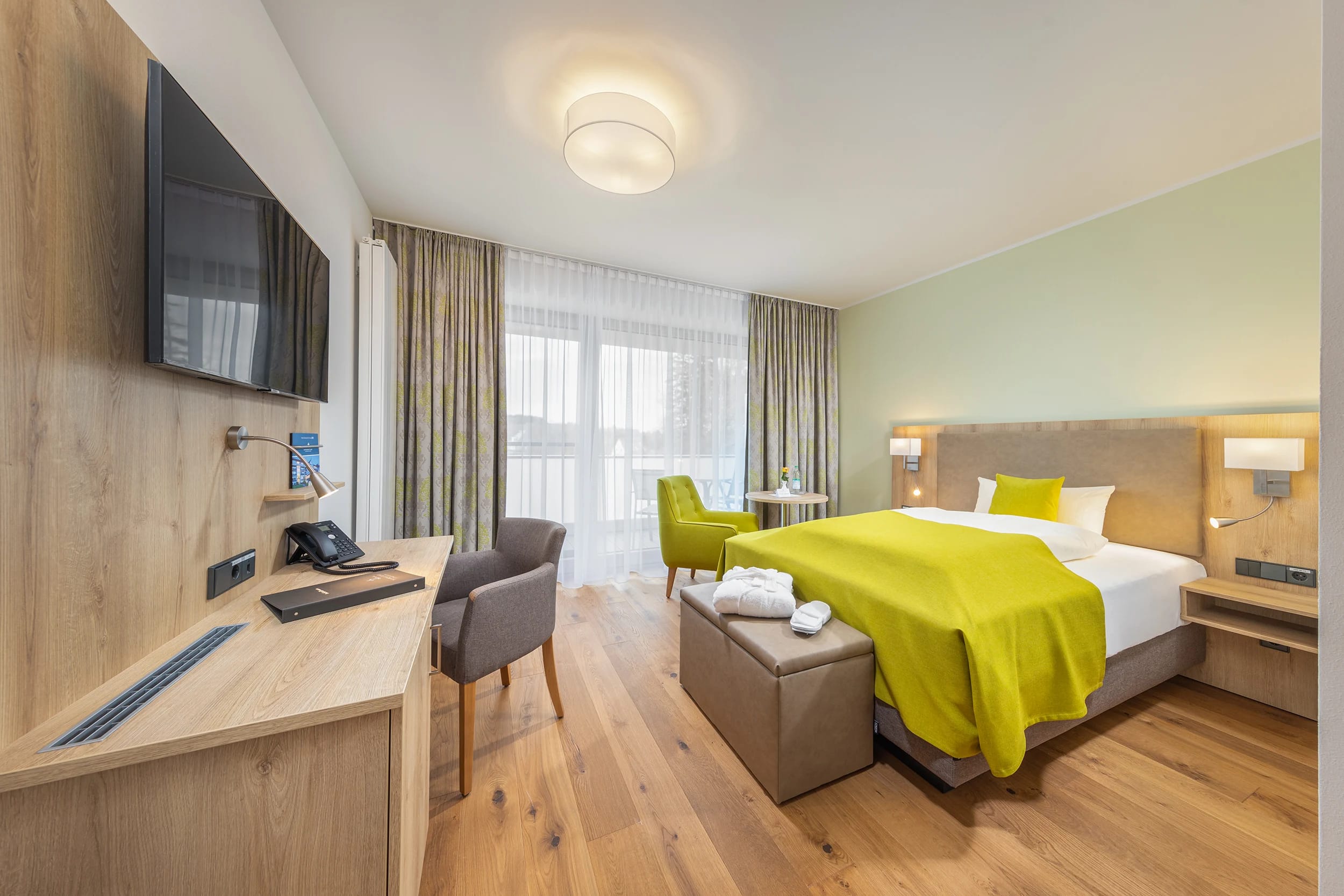 Zimmer mit Holzboden, Schreibtisch, Sessel und Bett mit grüner Tagesdecke