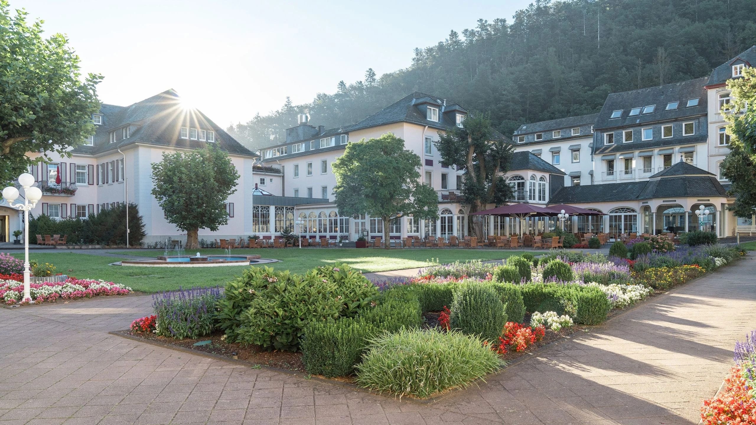 Außenansicht des Hotels mit Parkanlage, Blumenbeeten und bewaldetem Hang im Hintergrund