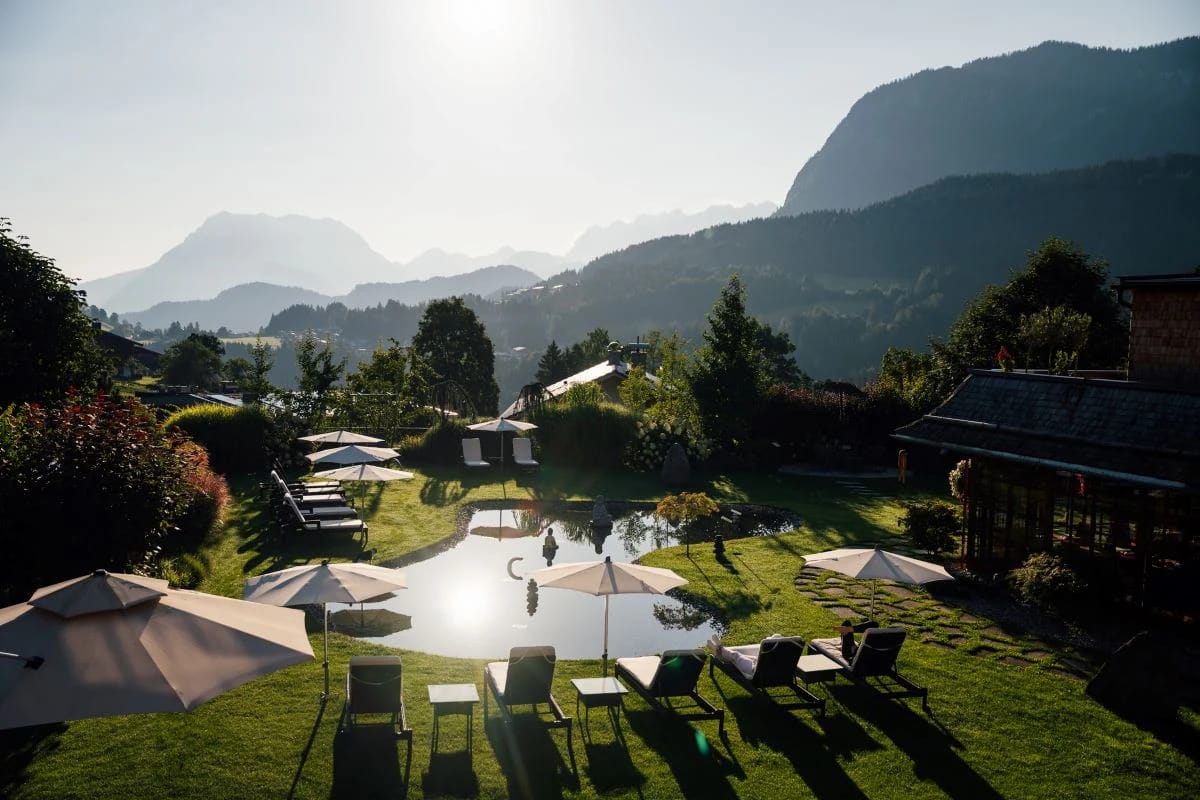 Naturbadeteich im Garten mit Liegestühlen und Sonnenschirmen vor Bergpanorama