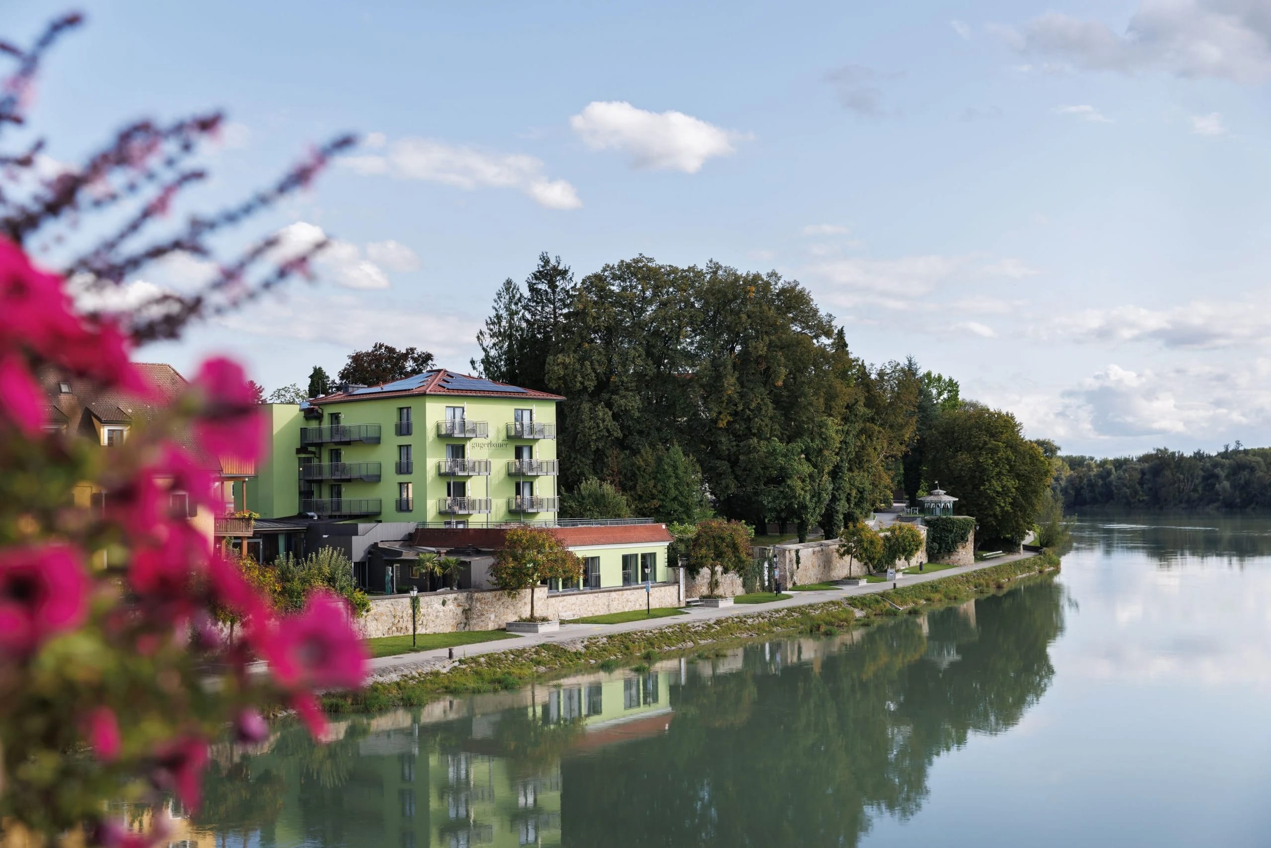 Grün gestrichenes Hotelgebäude direkt am Ufer des Inn, im Vordergrund blühende Sträucher
