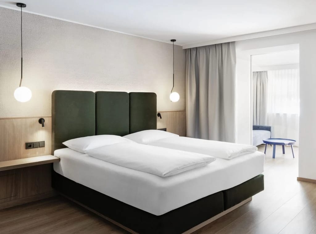 Doppelbett mit grünem Kopfteil und Pendelleuchten, dahinter ein angrenzender Wohnbereich
