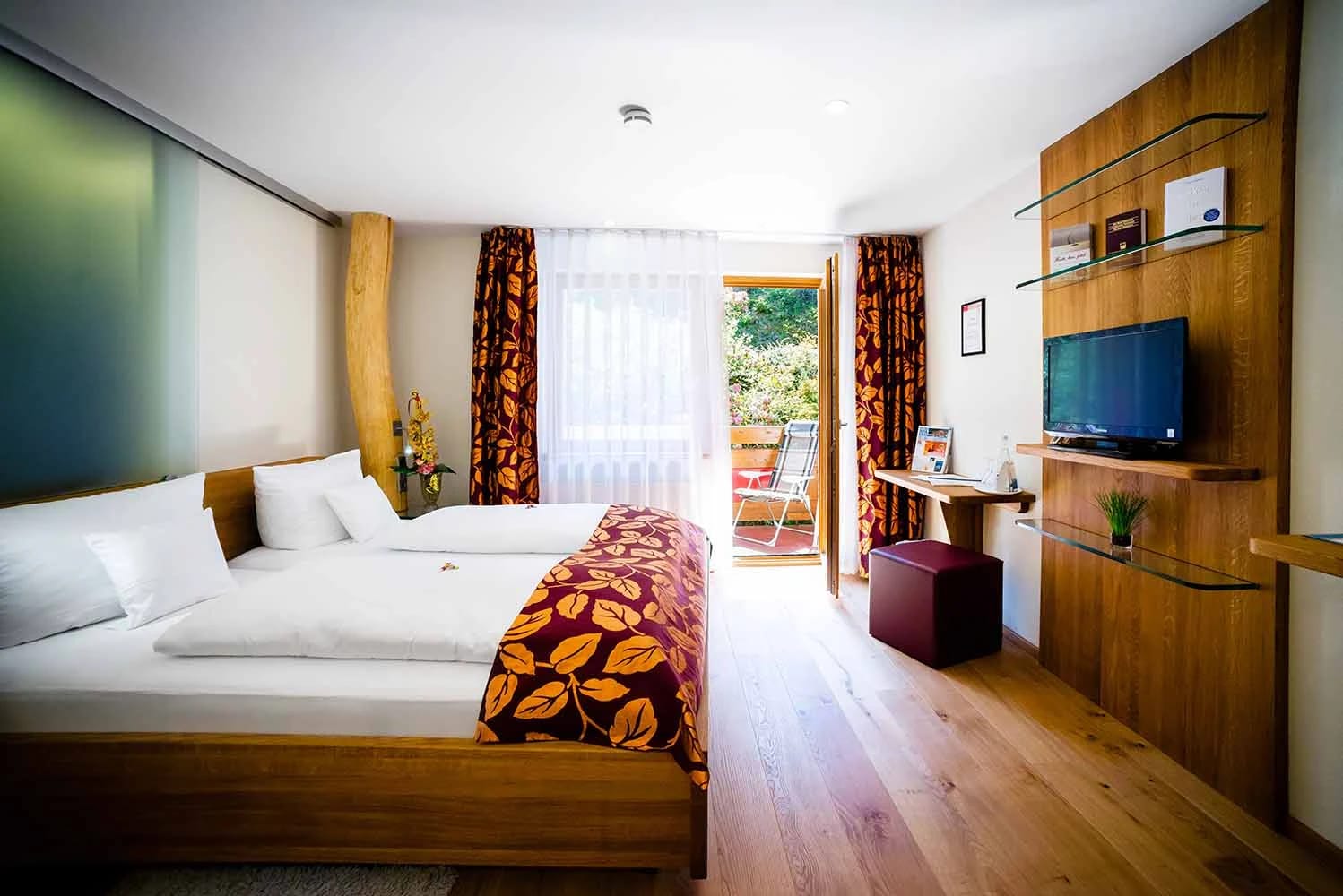 Zimmer mit Holzboden, Doppelbett mit blättergemusterter Tagesdecke, Schreibtisch mit Fernseher und Tür zum Balkon