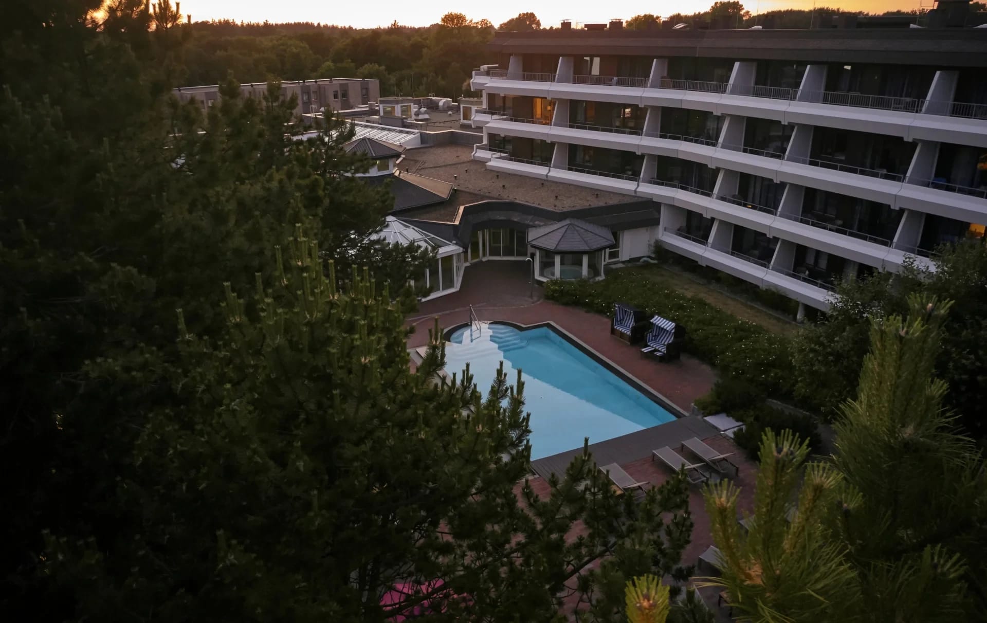 Blick von oben über den Außenpool auf das mehrstöckige Hotelgebäude mit Balkonen, umgeben von Bäumen