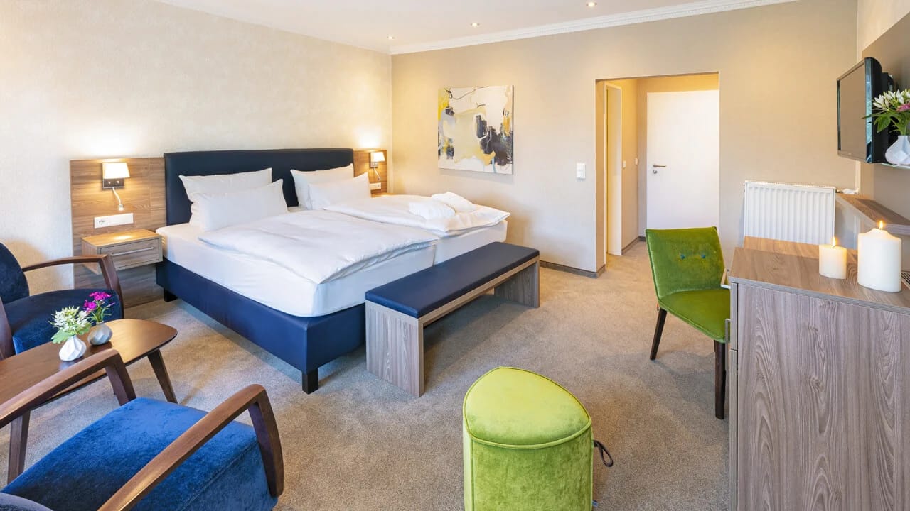 Geräumiges Doppelzimmer mit dunkelblauem Bettkopfteil, Sitzbank am Bettende, Sesseln und Schreibtisch am Fenster