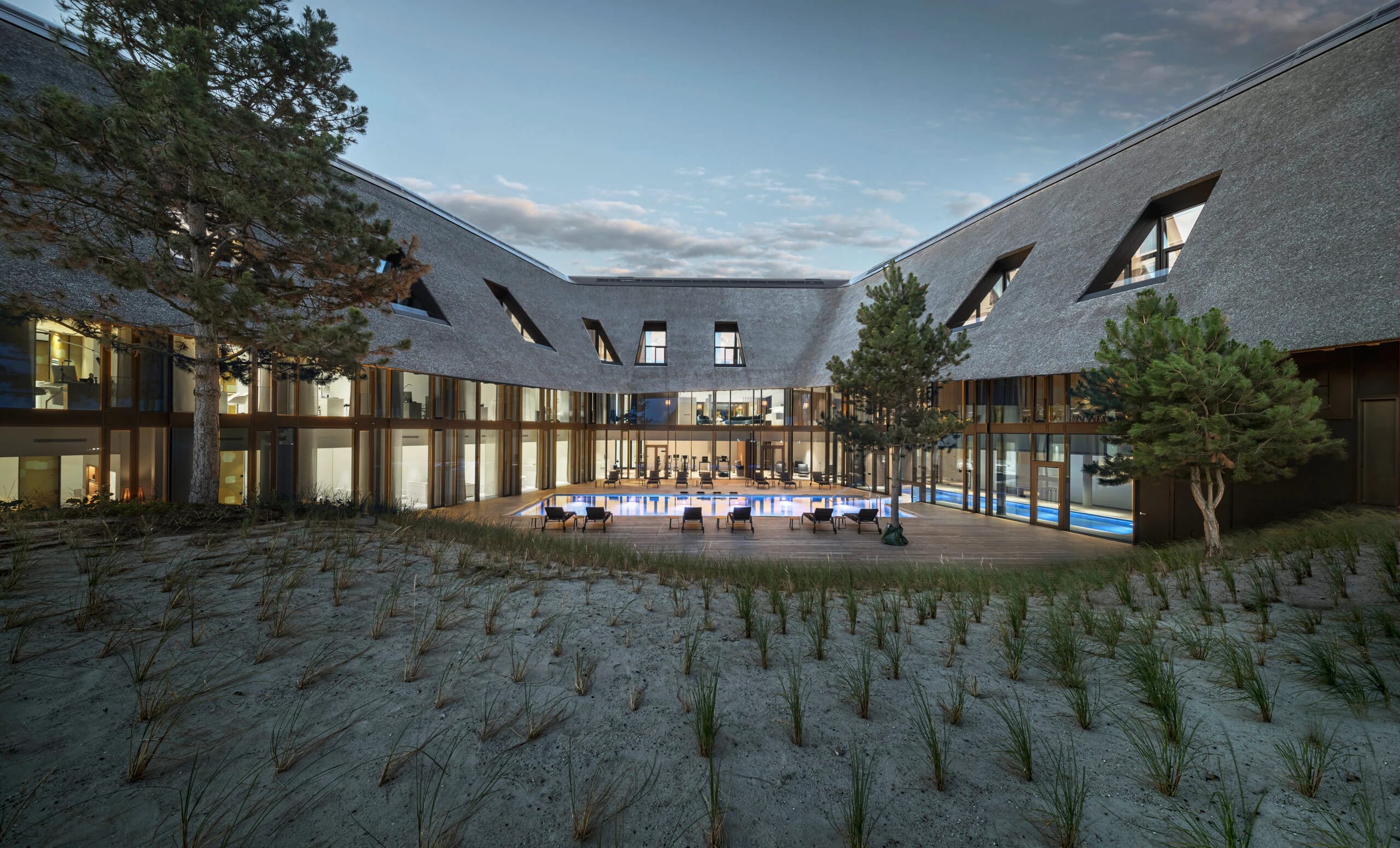 Innenhof zwischen den reetgedeckten Gebäudeflügeln mit Holzdeck, langer Tafel und Pool in der Dämmerung
