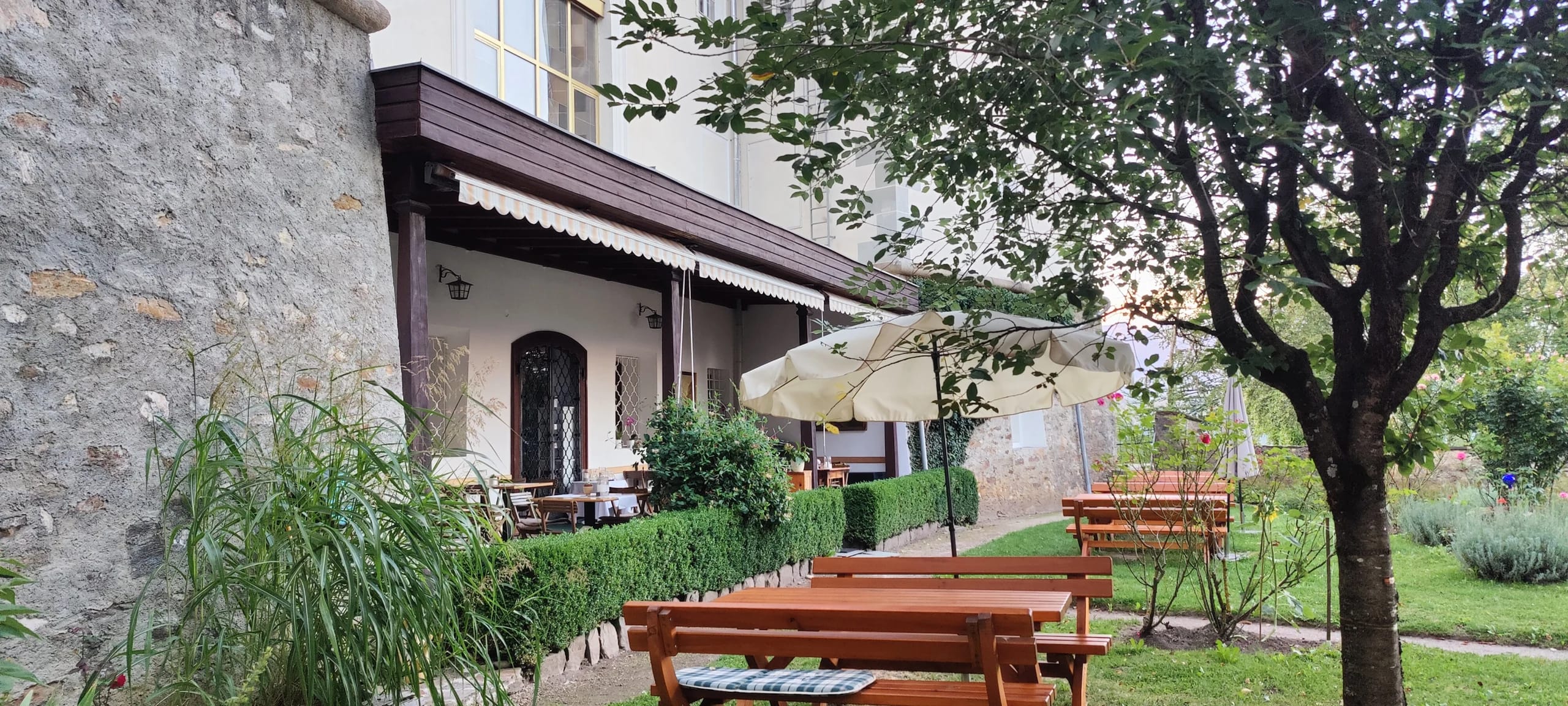 Terrasse mit Holzbänken und Sonnenschirm entlang der Hausmauer, daneben Bäume und Sträucher