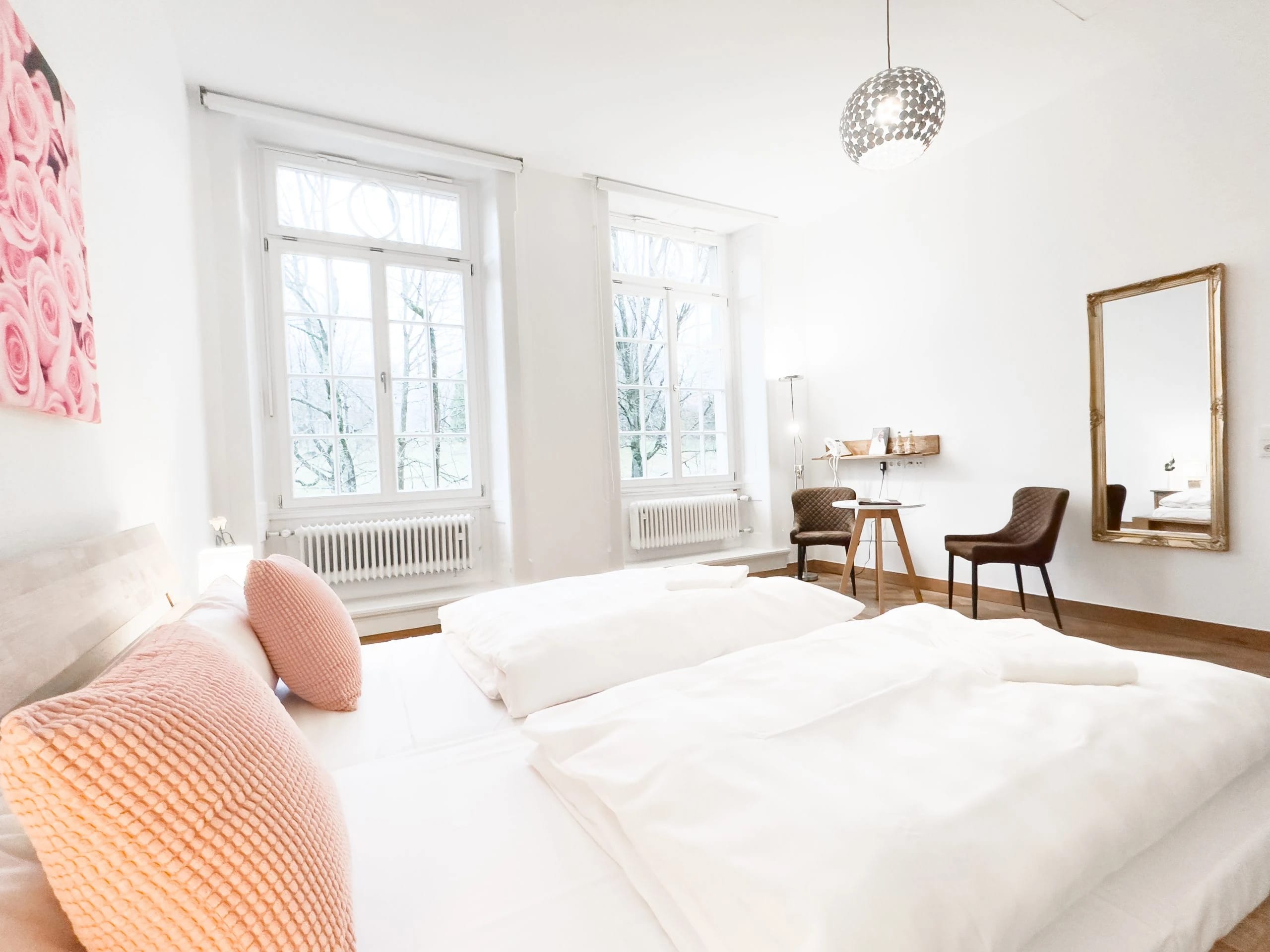 Helles Gästezimmer mit Doppelbett, hohen Sprossenfenstern und goldgerahmtem Spiegel
