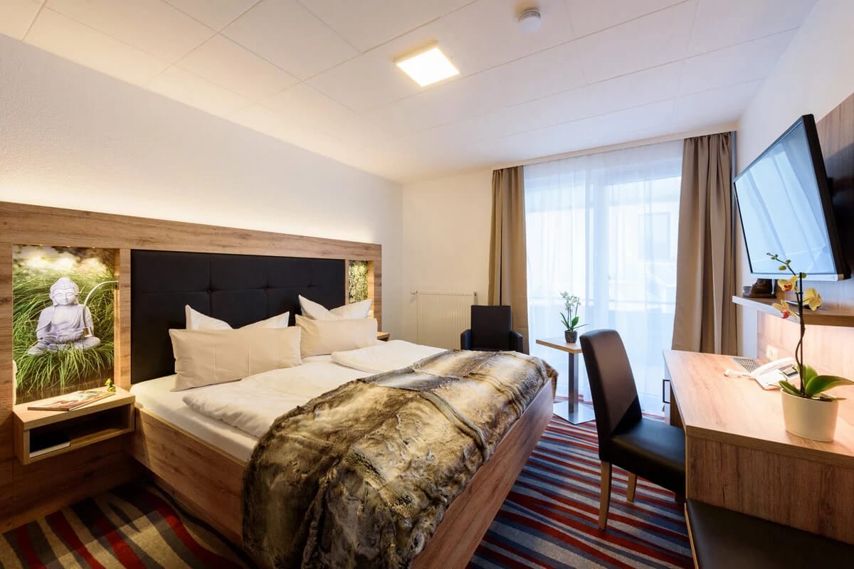 Hotelzimmer mit Doppelbett, Fellplaid, beleuchtetem Bild am Kopfende, Schreibtisch und Wandfernseher