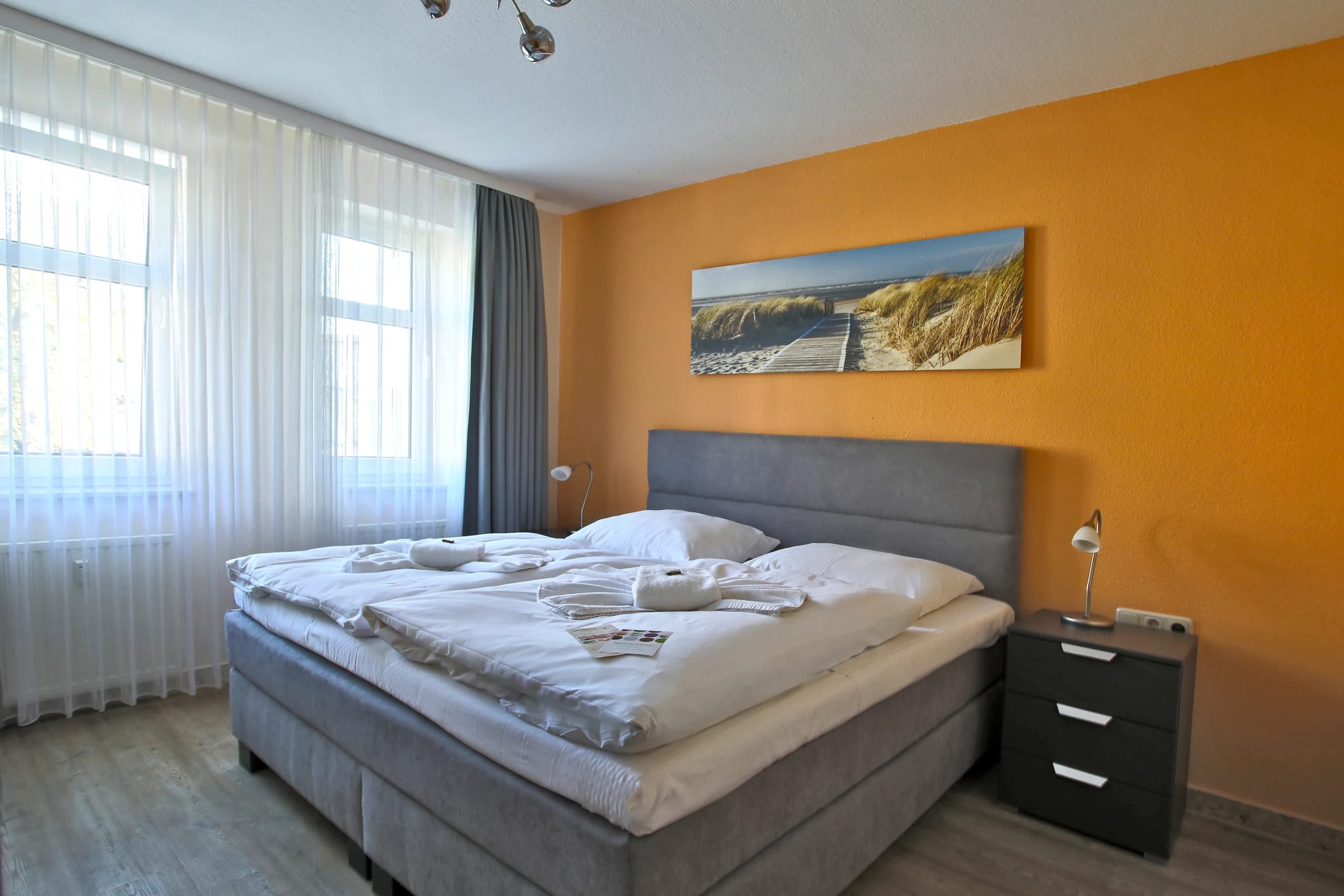 Doppelzimmer mit Boxspringbett vor orangefarbener Wand, Dünenbild über dem Bett und zwei Fenstern