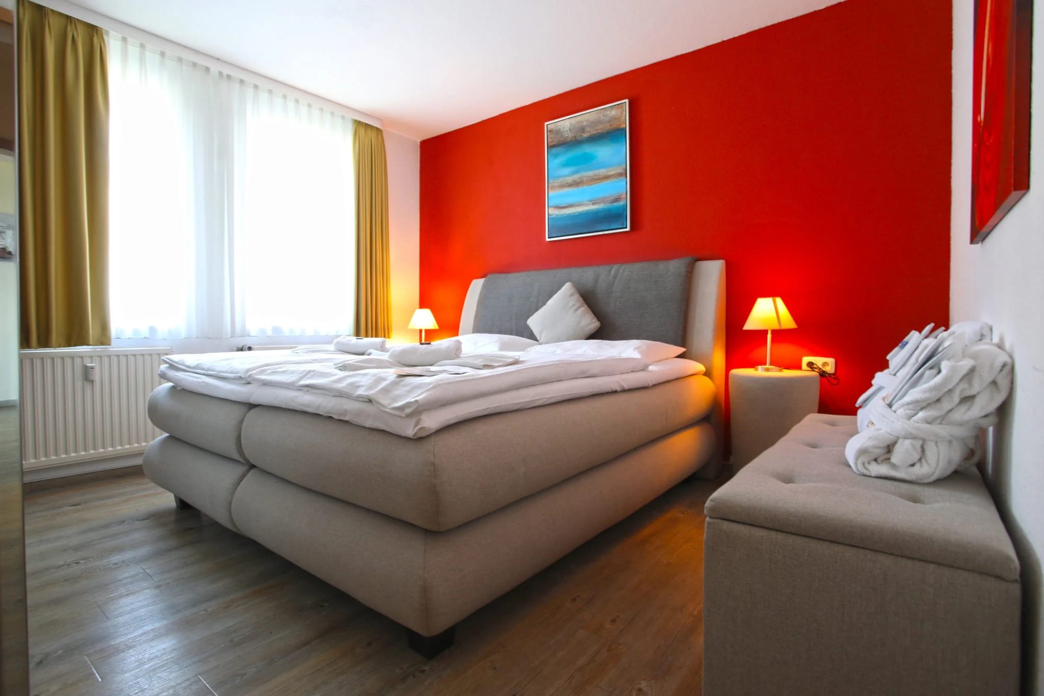 Doppelzimmer mit Boxspringbett vor roter Wand, Nachttischlampen und gepolsterter Bank am Fußende