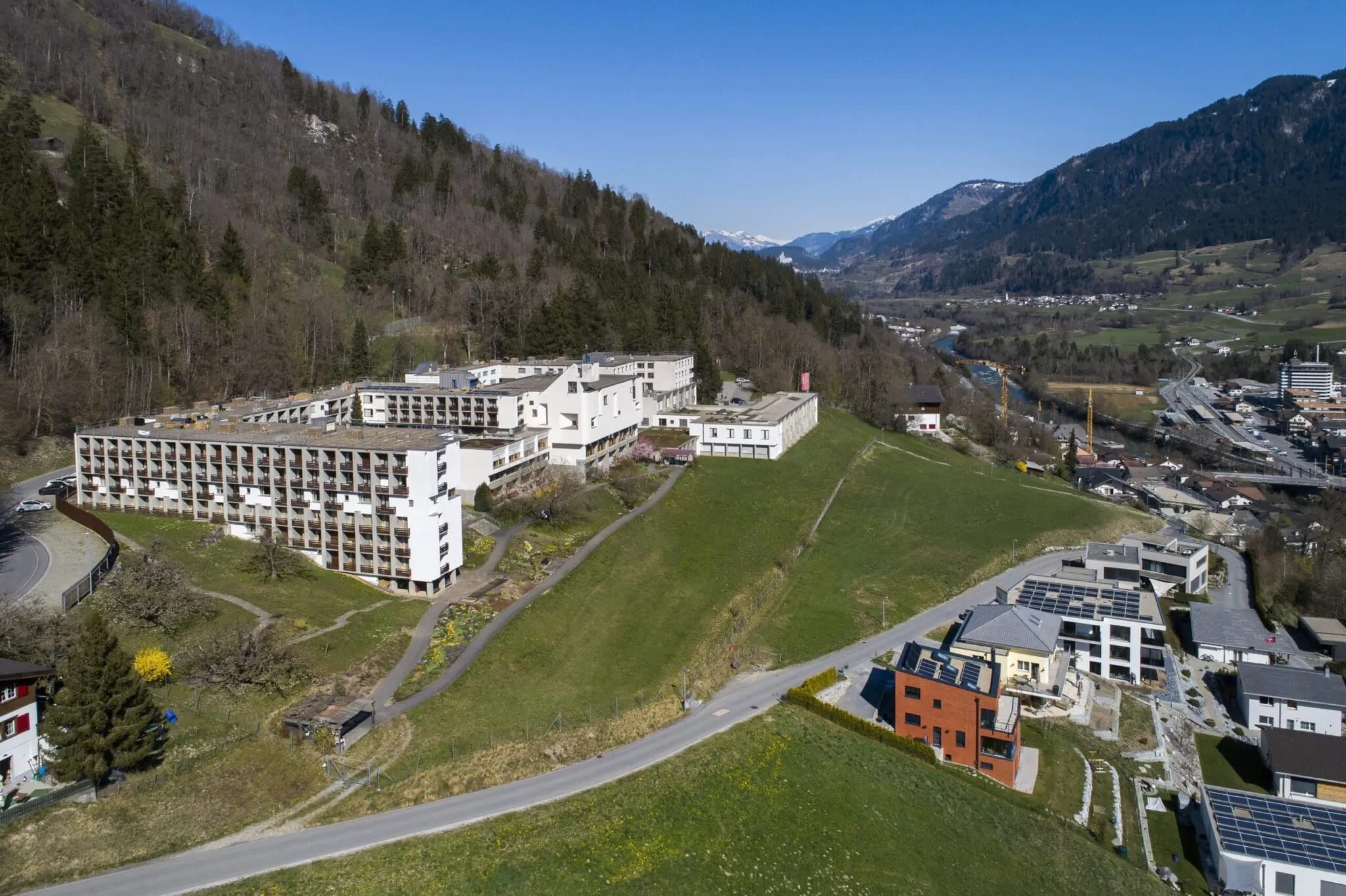 Luftaufnahme der weißen Klosteranlage auf dem Hang über dem Tal, dahinter Berge