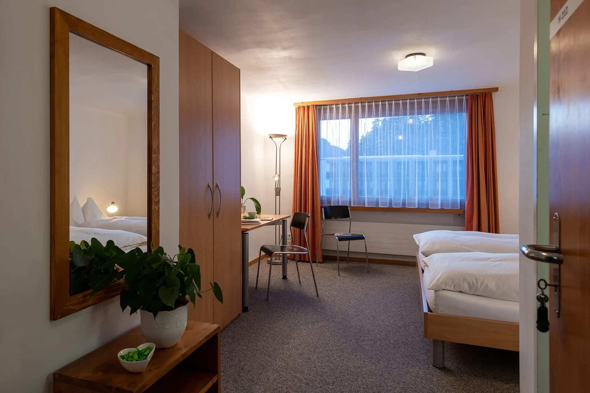 Gästezimmer mit Bett, Schreibtisch, Spiegel, Schrank und Fenster mit orangefarbenen Vorhängen