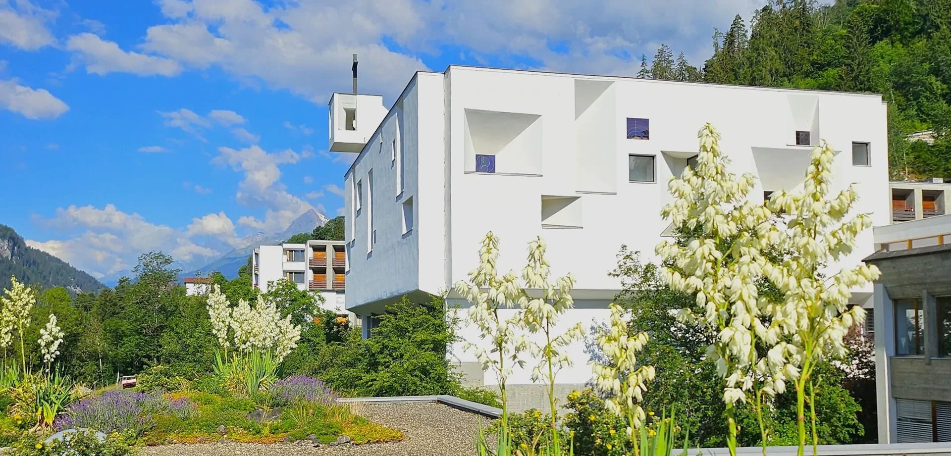 Weiße Fassade des Klostergebäudes mit blühendem Garten und Bergen im Hintergrund