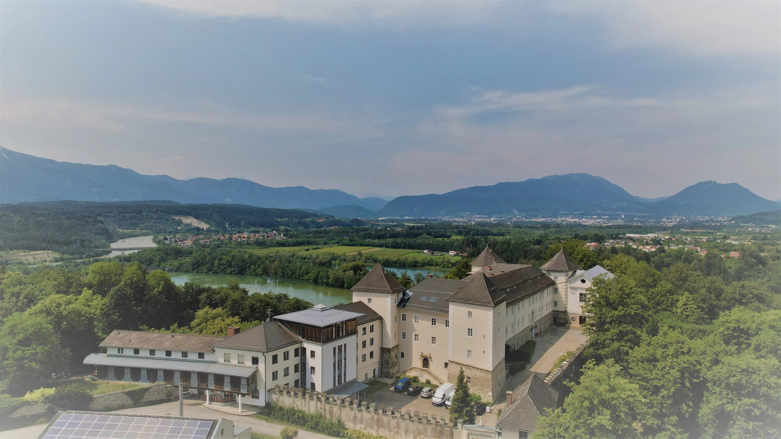 Luftaufnahme der Klosteranlage mit Turm und Erkertürmchen, Wiesen, Fluss im Tal und Bergen im Hintergrund