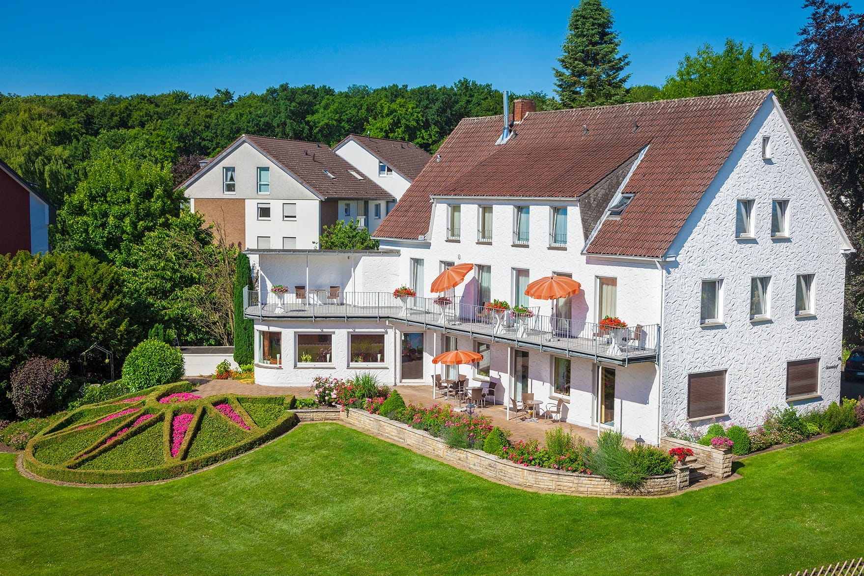 Weißes Gästehaus mit Balkon und orangefarbenen Sonnenschirmen, davor eine Rasenfläche mit bepflanztem Blumenbeet