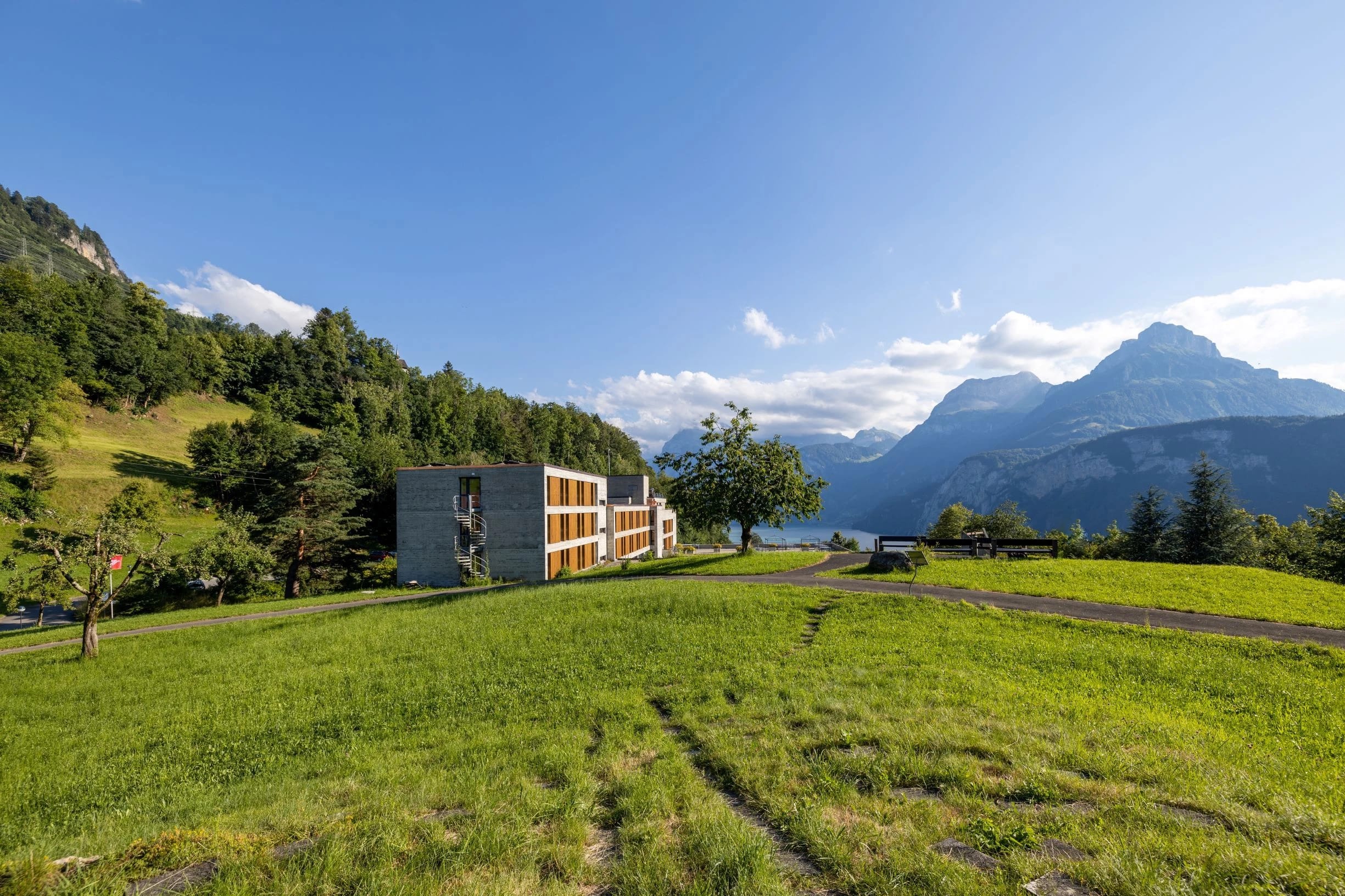 Langgestrecktes Haus mit Holzlamellen-Fassade auf einer Sommerwiese, dahinter Berge und See