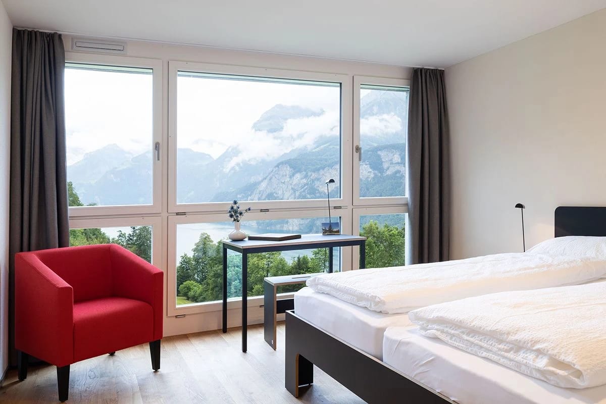 Zimmer mit Panoramafenstern und Blick auf schneebedeckte Berge, rotem Sessel und Schreibtisch