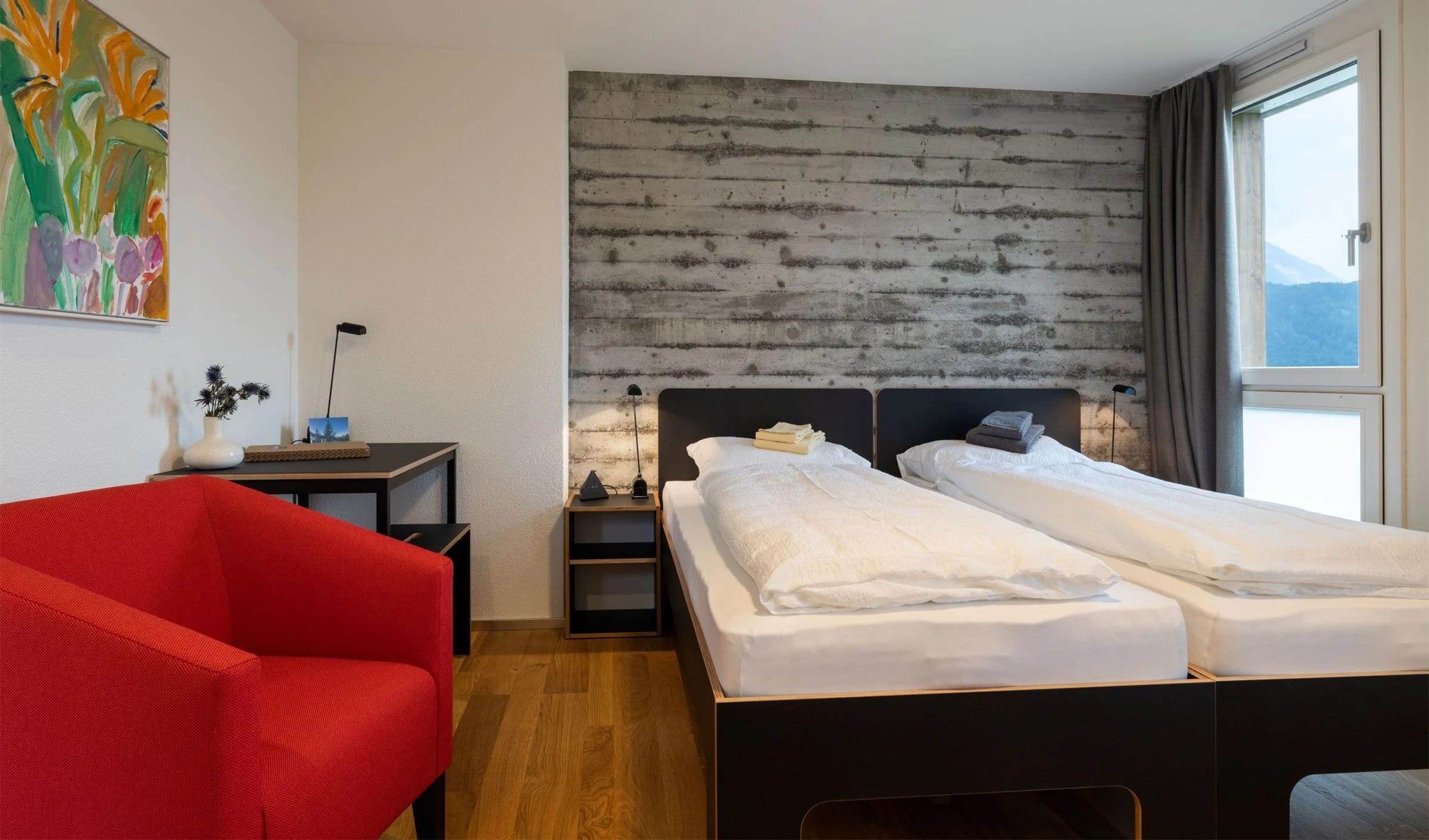 Zimmer mit zwei zusammengestellten Betten vor Sichtbetonwand, rotem Sessel und Fenster