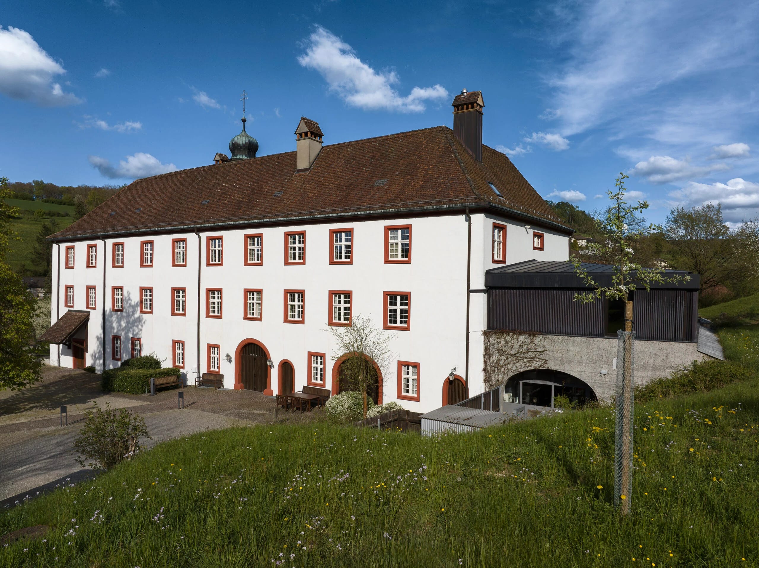 Fassade des Hauptgebäudes mit rot gefassten Fenstern, davor eine blühende Wiese
