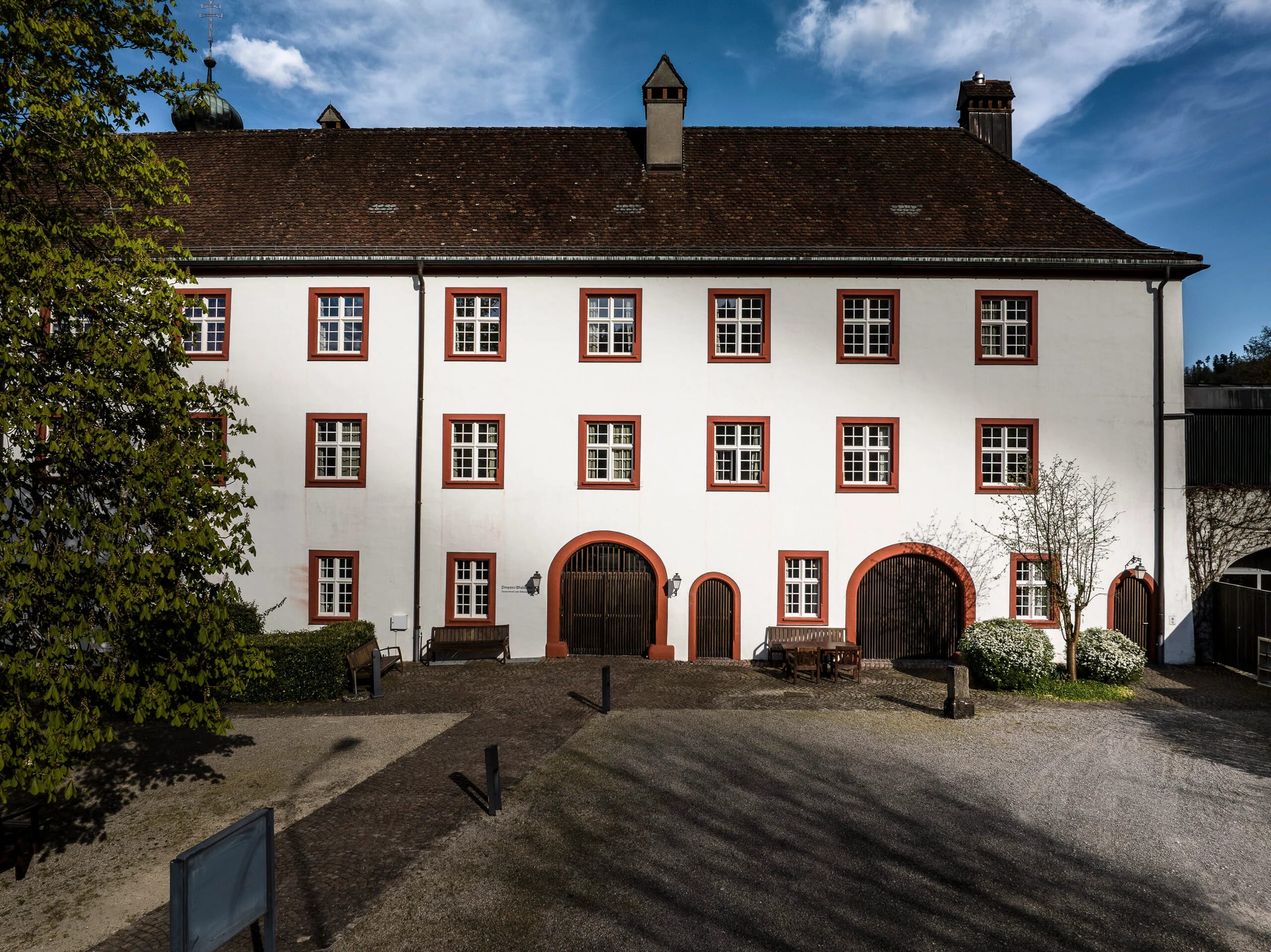 Innenhoffassade mit rot gerahmten Fenstern, Rundbogentoren und Sitzbänken