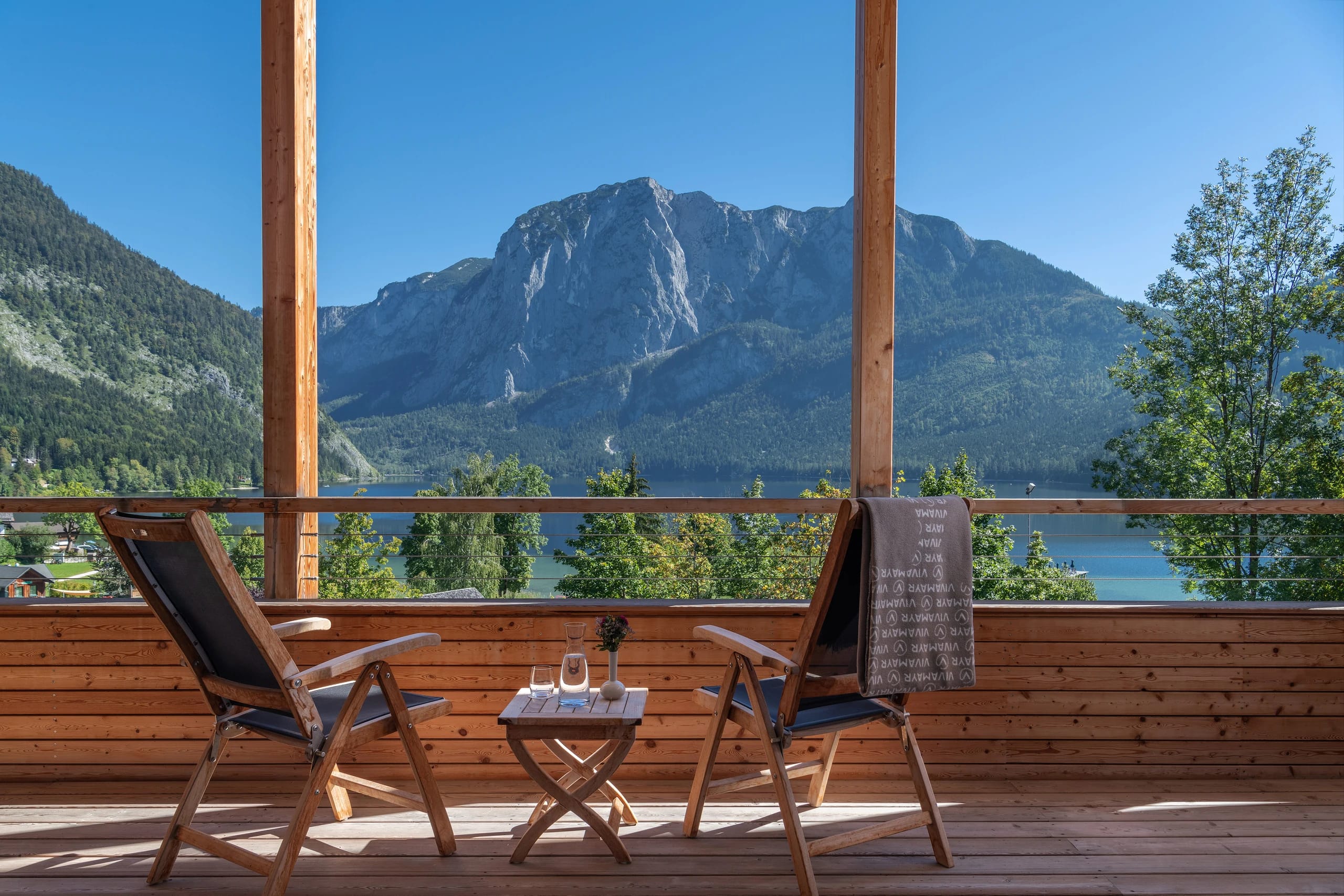Holzbalkon mit zwei Liegestühlen und kleinem Tisch, Blick auf Bergmassiv und See