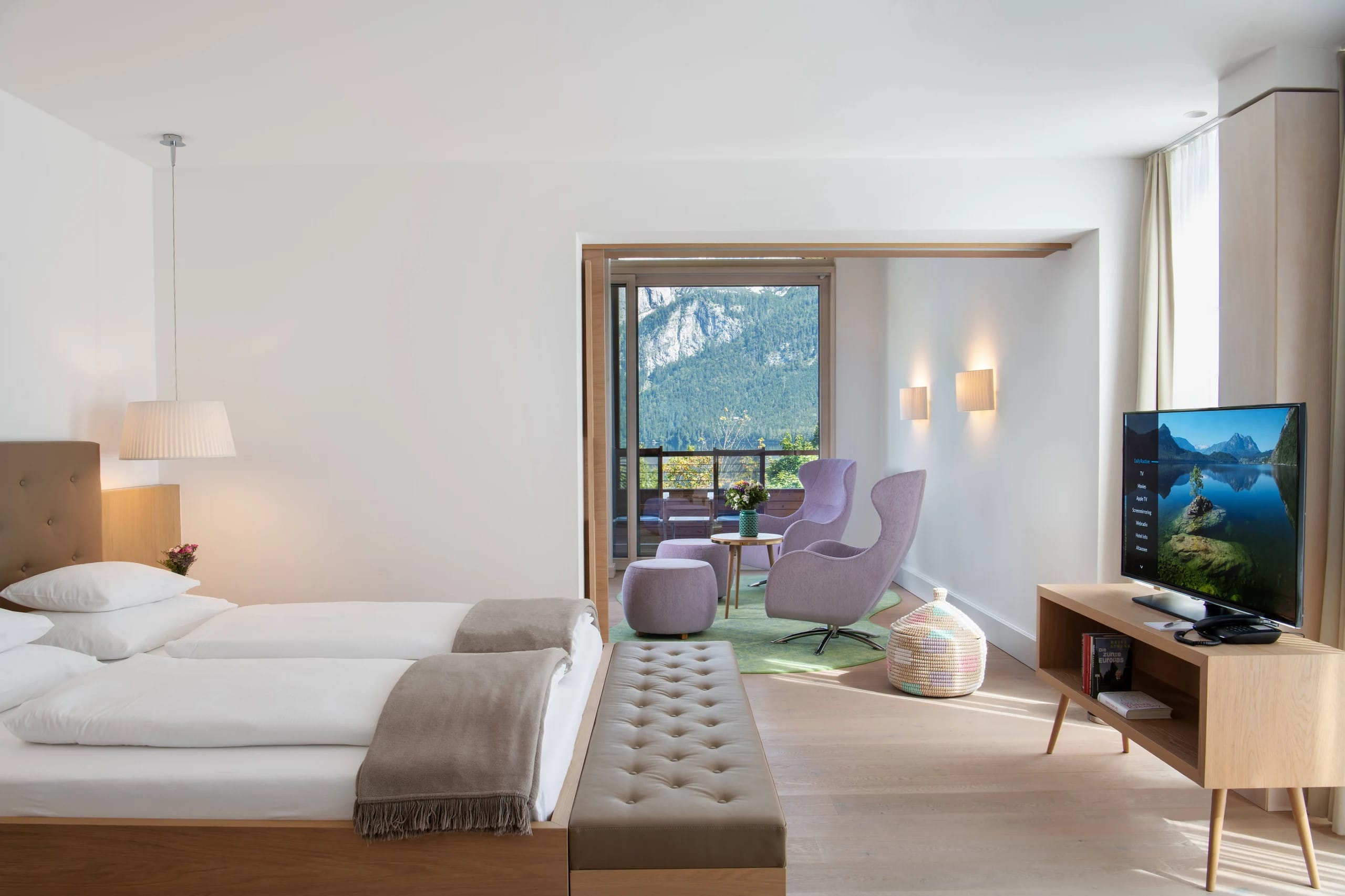 Helles Zimmer mit Doppelbett, Sesseln und offener Tür zum Balkon mit Bergblick