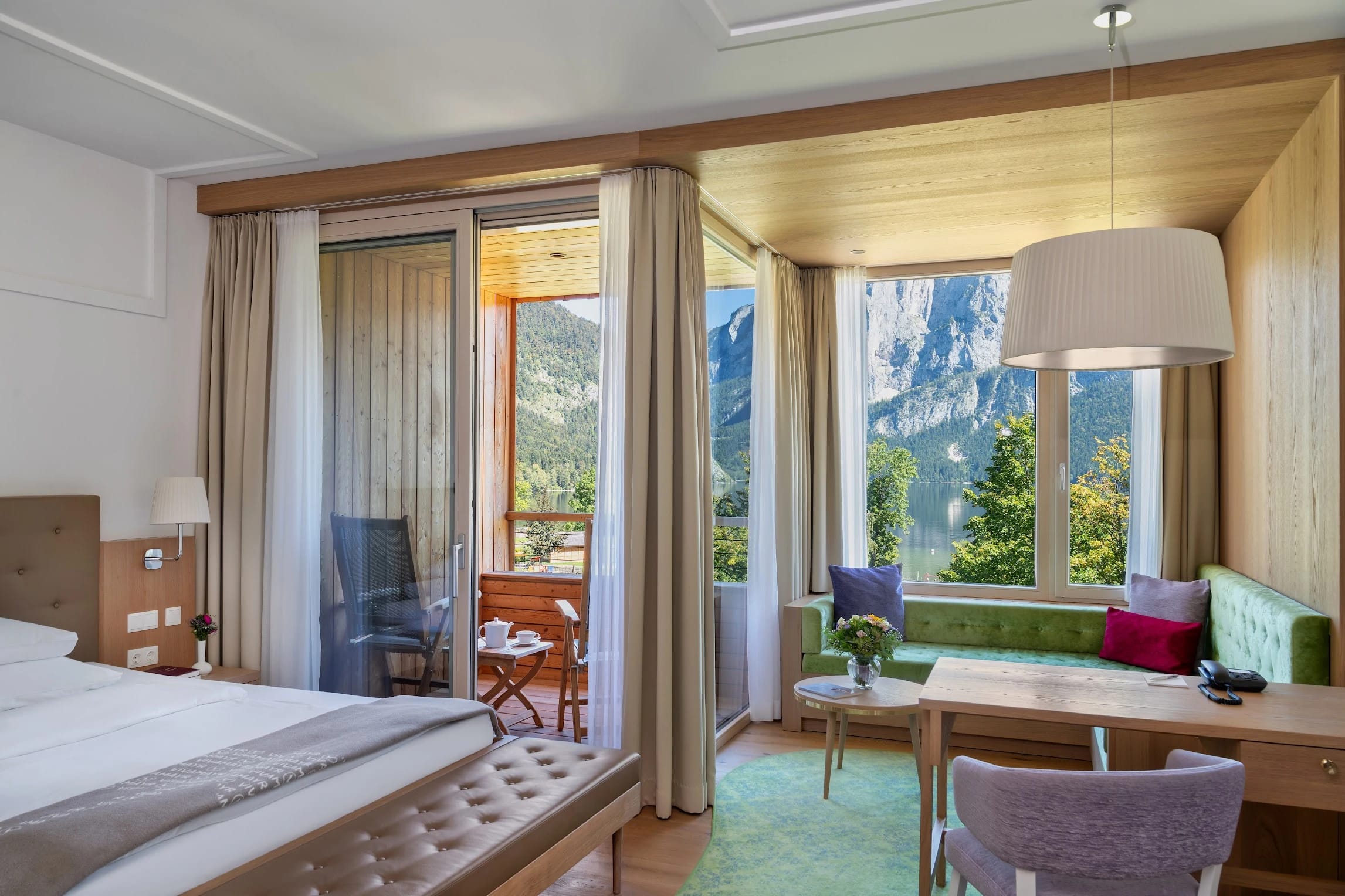 Zimmer mit Doppelbett, Schreibtisch, gepolsterter Fensterbank und Balkon mit Blick auf Berge und See