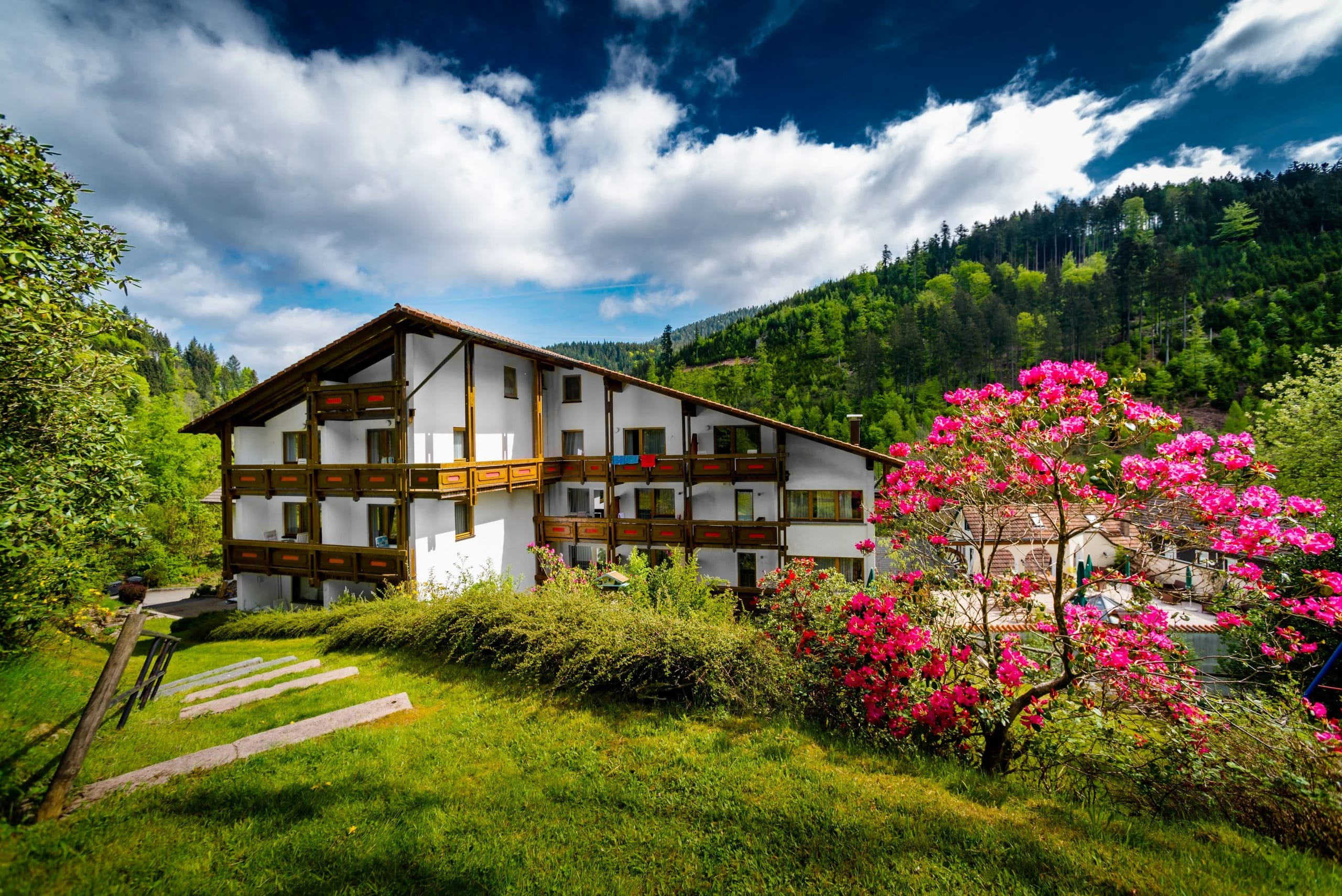 Weiß-braunes Hausteil mit Holzbalkonen auf einer Wiese, daneben ein blühender Rhododendron