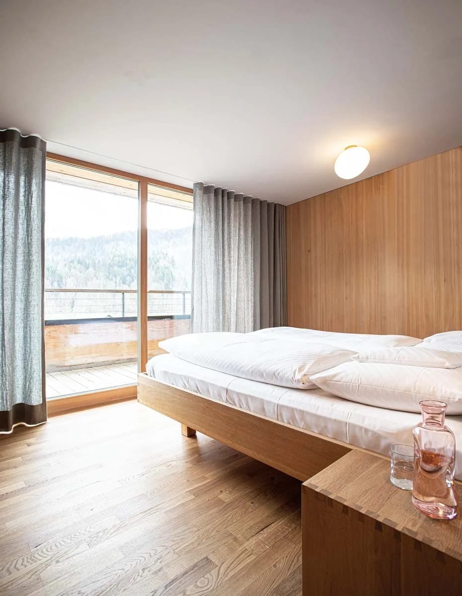 Zimmer mit Doppelbett vor Holzwand und bodentiefem Fenster mit Balkon und Blick auf den Wald