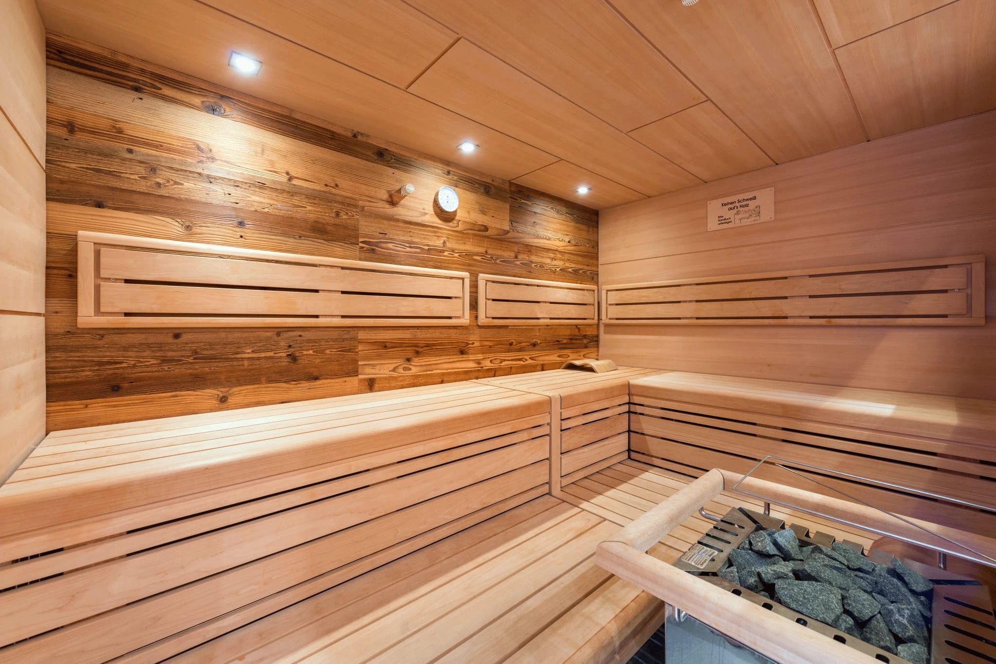 Sauna mit hellen Holzbänken, Saunaofen mit Steinen und Deckenstrahlern