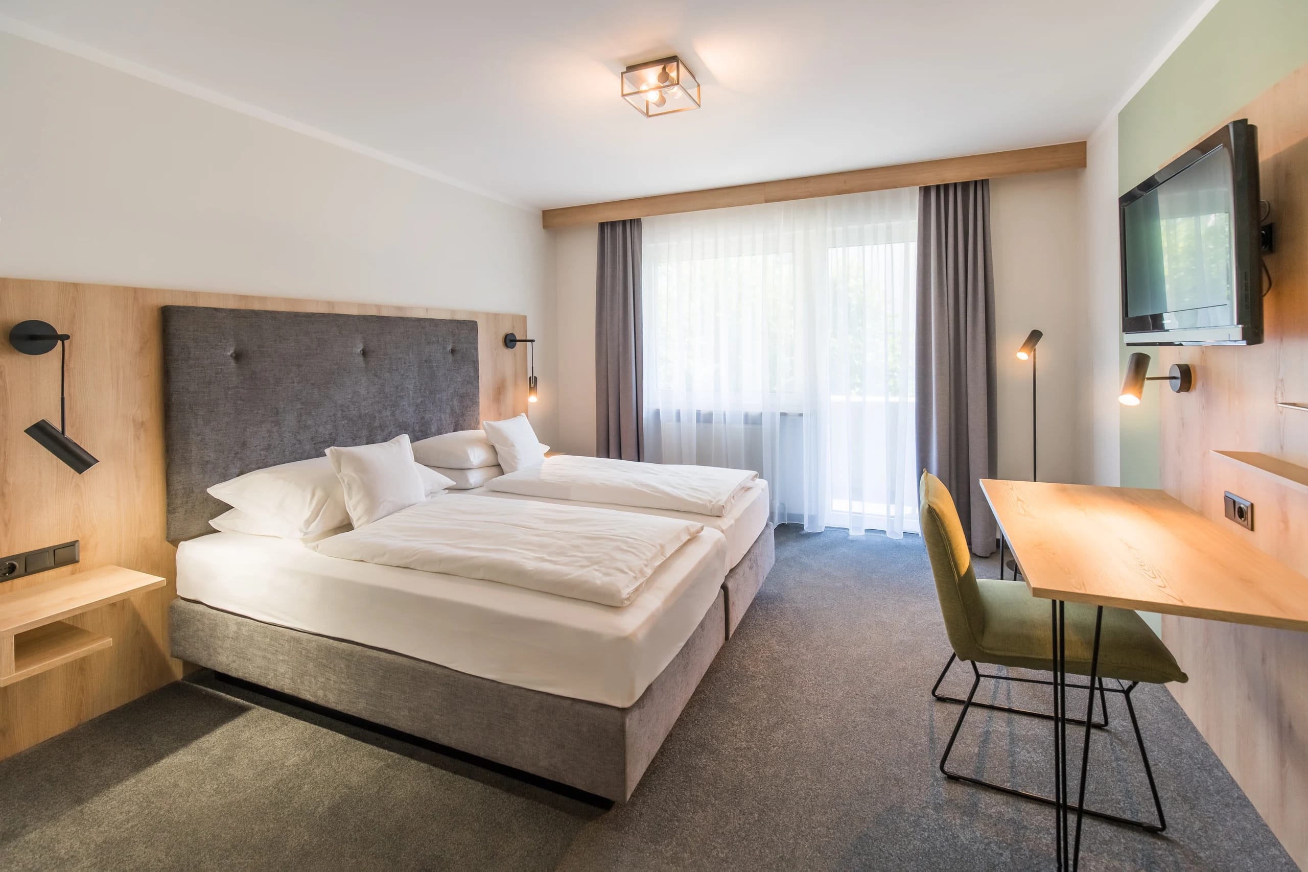 Doppelzimmer mit Doppelbett, gepolstertem Kopfteil aus Holz, Schreibtisch mit Stuhl und Fenster mit Vorhängen