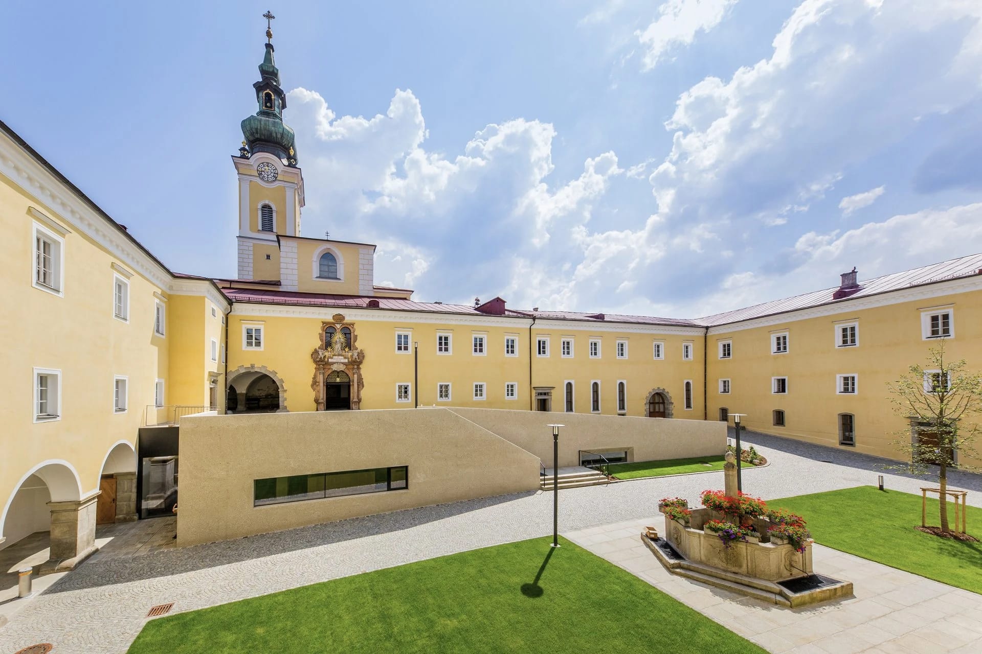 Innenhof des Stifts mit gelben Klostergebäuden, Kirchturm und Rasenflächen unter blauem Himmel