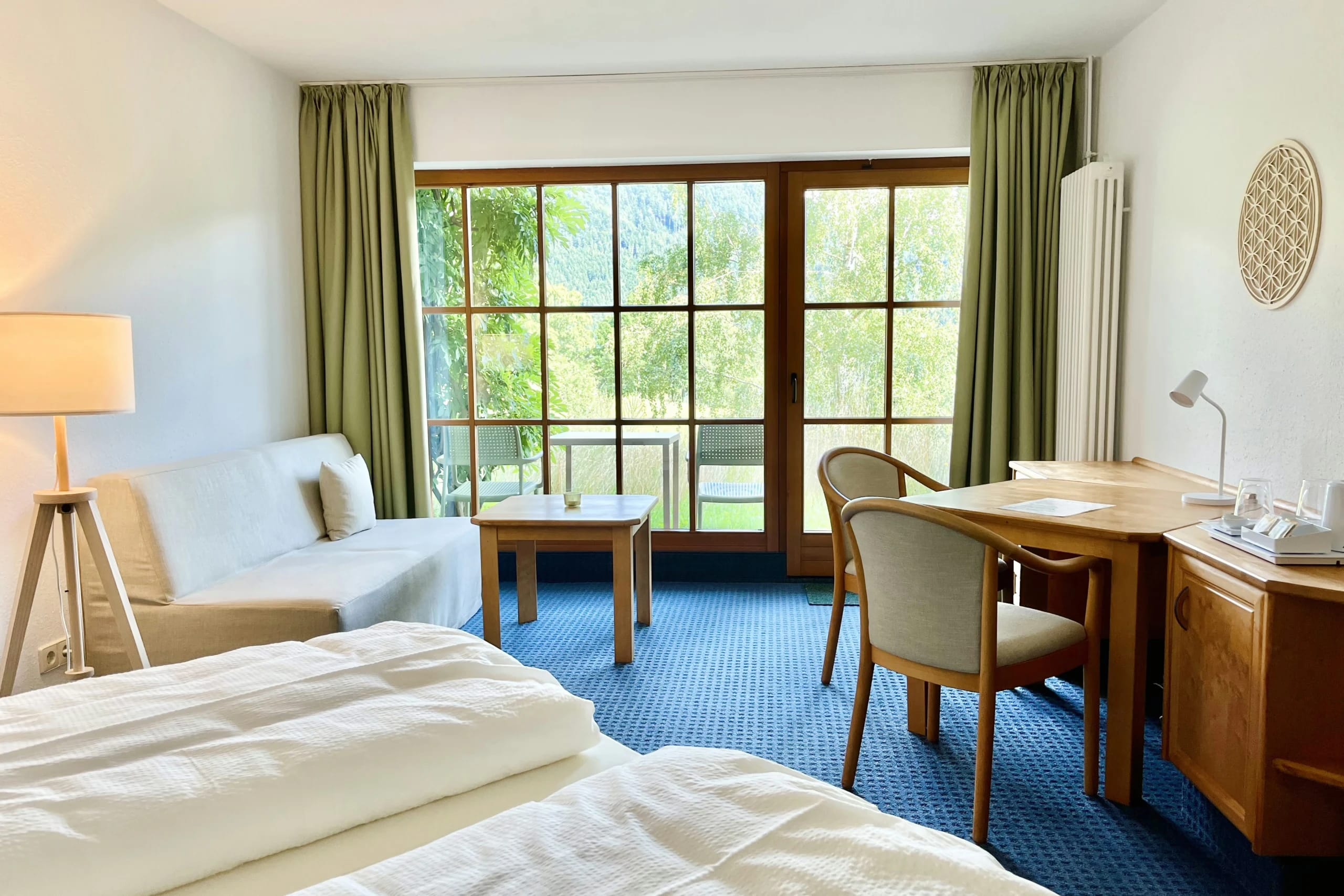 Helles Zimmer mit Bett, weißem Sofa, Schreibtisch und Sprossentür zum Balkon, grüne Vorhänge