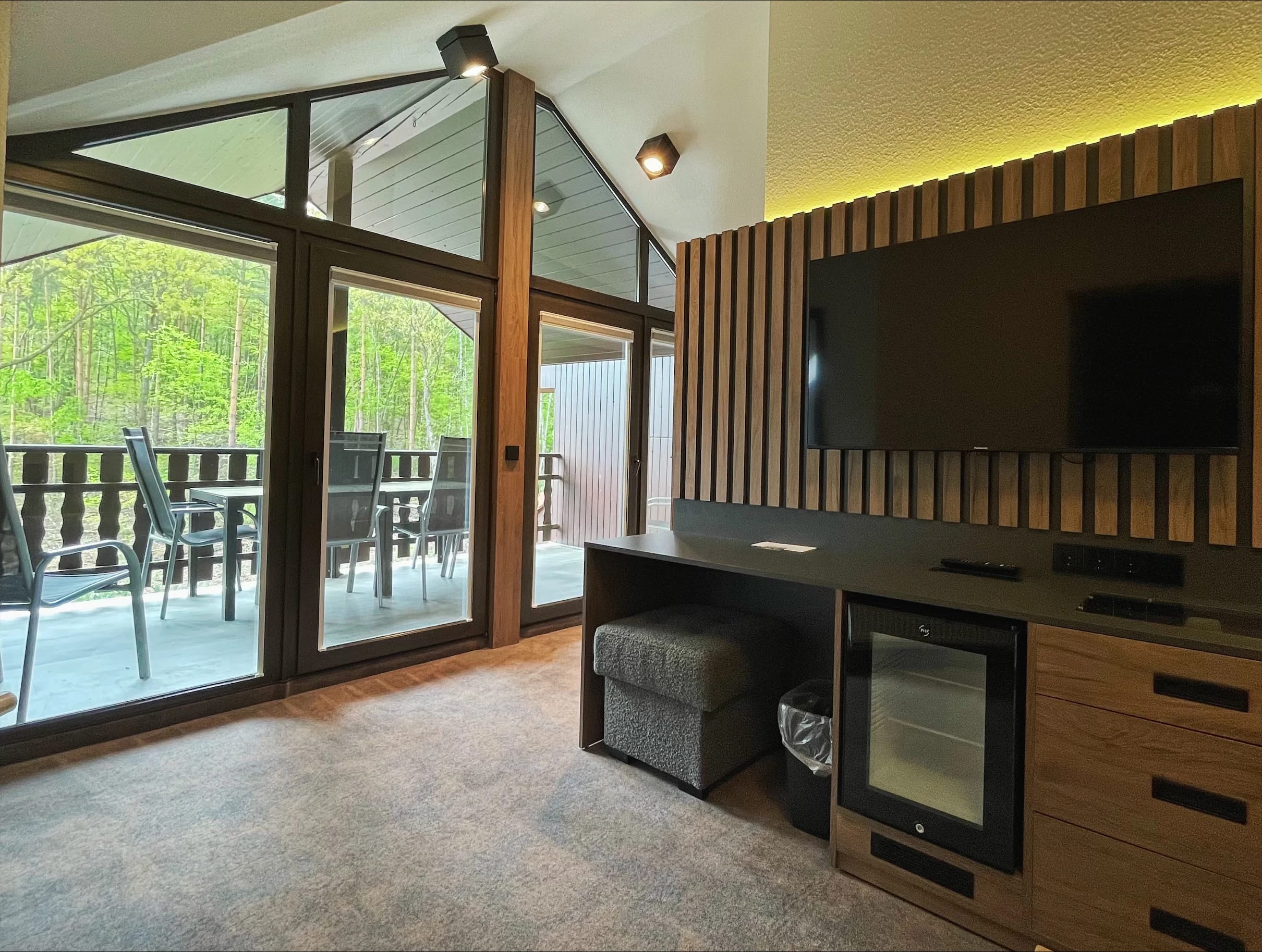 Zimmer mit Sideboard, Fernseher an der Holzlamellenwand und Glastür zum Balkon mit Blick ins Grüne