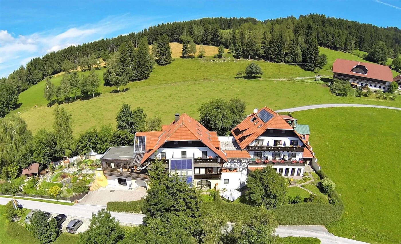 Luftaufnahme des Hauses auf einem grünen Hang vor bewaldeten Hügeln