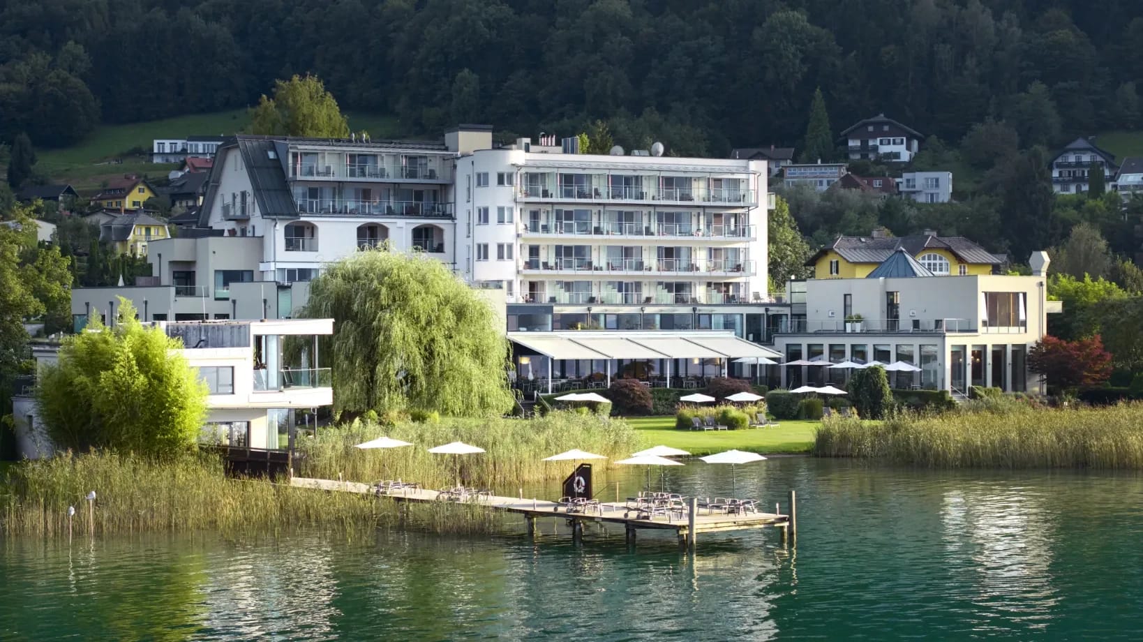 Blick vom Wörthersee auf das weiße Haupthaus mit Balkonen, davor Liegewiese, Sonnenschirme und ein Badesteg