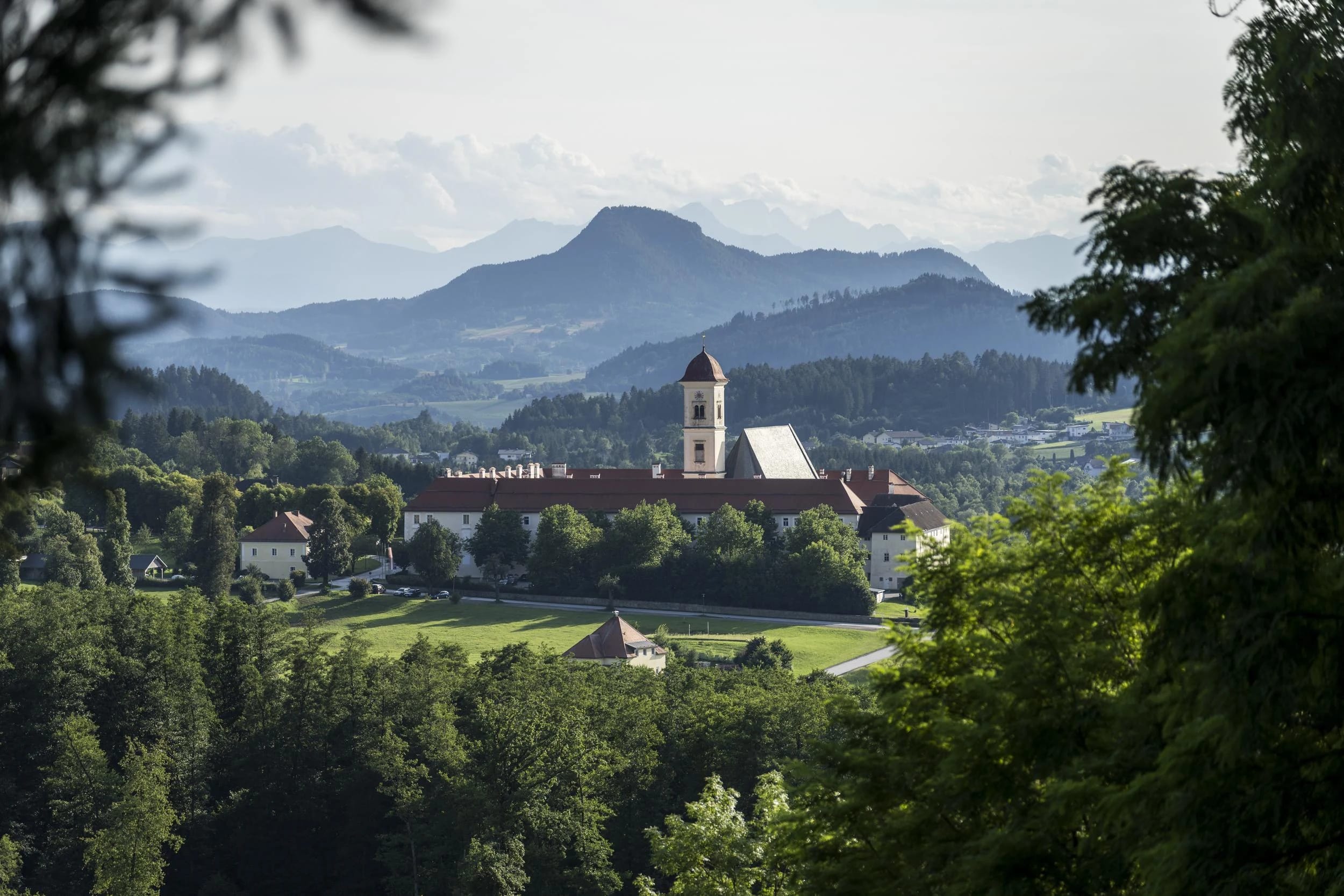 Blick über Wiesen und Wald auf das Stift mit Kirchturm, dahinter bewaldete Hügel und Berge
