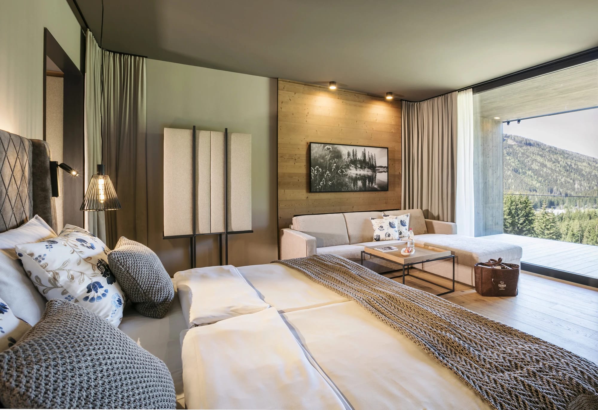 Hotelzimmer mit Doppelbett, Sofa, Couchtisch und großem Fenster mit Blick auf bewaldete Hügel