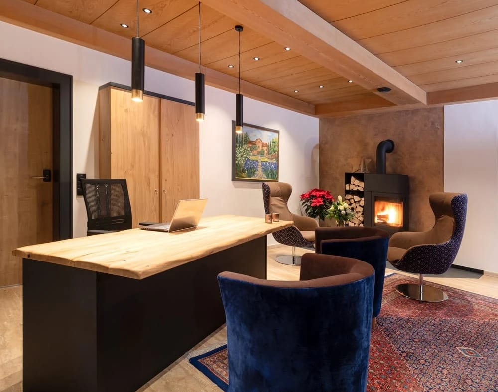 Rezeptionstresen aus Holz mit Pendelleuchten, davor Sessel vor einem brennenden Kaminofen