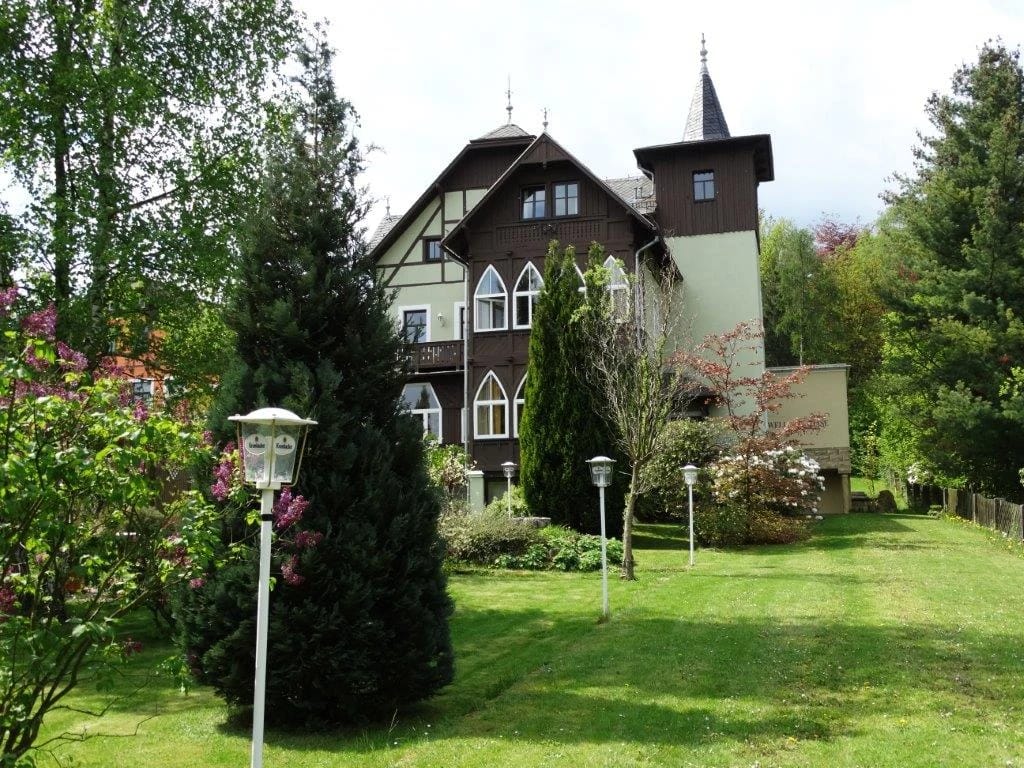 Dunkel verschaltes Haus mit Turm hinter Rasenfläche, Sträuchern und weißen Wegleuchten